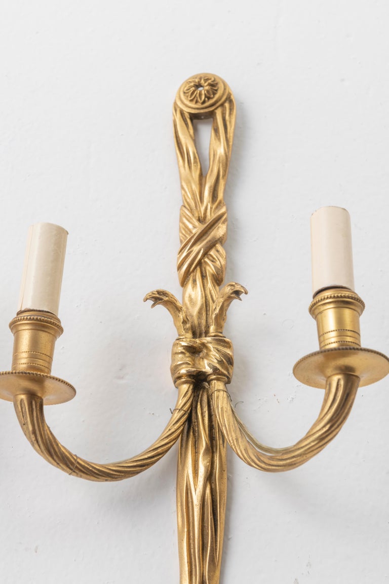 Lovely pair of gilt bronze sconces in the shape of knotted ribbons holding two candle arms, produced in 19th century France.

Each sconce features tassels, draped over buttons, decorated with rosettes. The neoclassical elements, typical of the