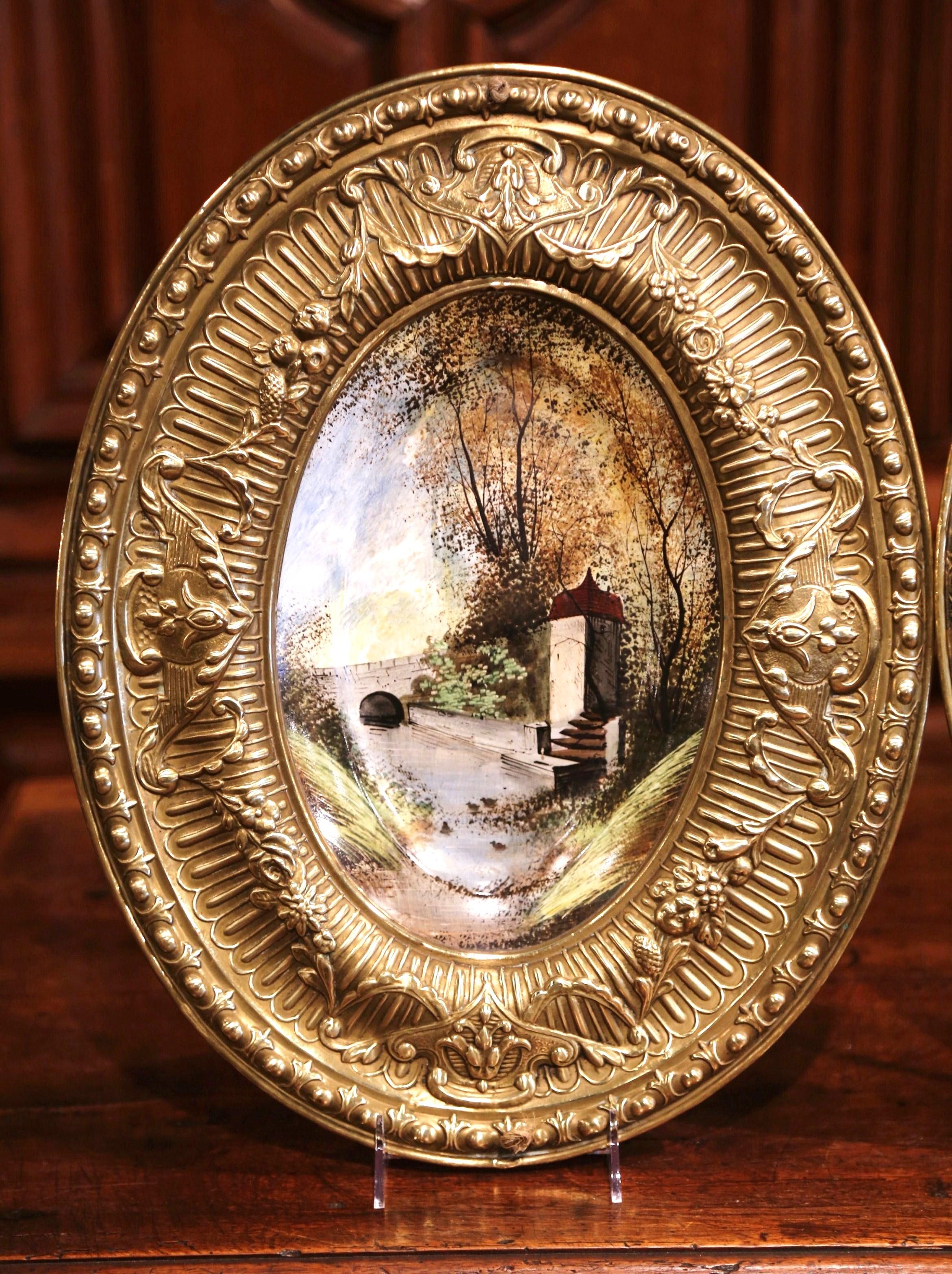 Decorate a den or game room with this pair of antique oval wall plaques. Crated in France circa 1870, each porcelain composition is set inside a repousse brass frame. The ceramic plaques feature hand painted landscape scenes with chateau, trees,