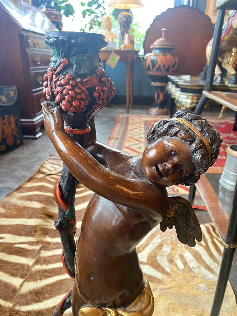 A pair of large identical patinated and polychrome Bronze Cherubs in the NeoClassical style. Both stand atop of a platform base while upholding grape adorned vases.