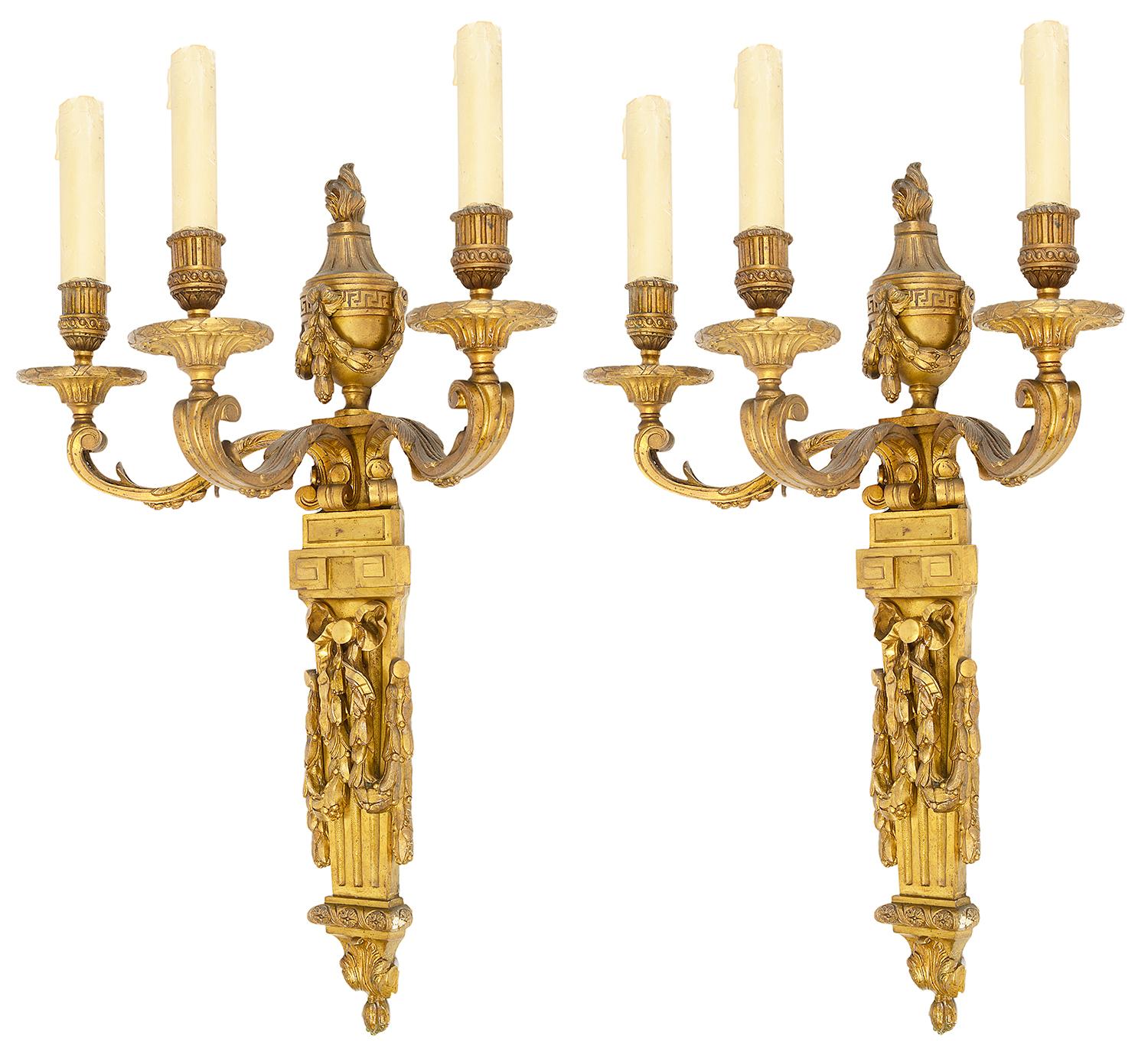 An impressive pair of 19th century gilded ormolu three branch wall sconces, each with flaming urns, ribbon and swag decoration over tapering fluted back plates.