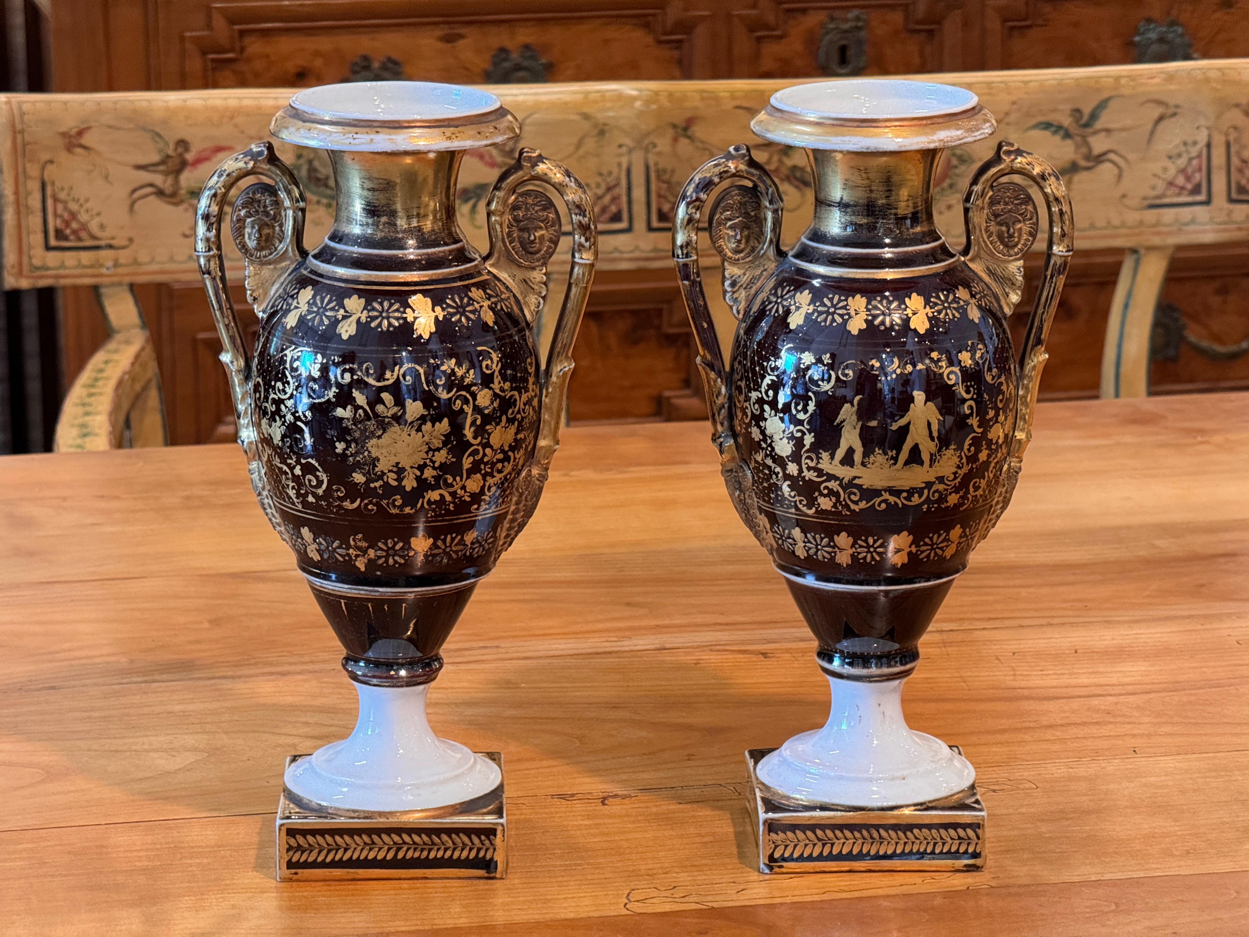 A pair of 19th century Old Paris porcelain urns featuring classical forms with double scroll handles and square bases. Finished in a deep cobalt ground with gilt neoclassical figures and floral motifs, the urns retain vibrant hand-applied detailing