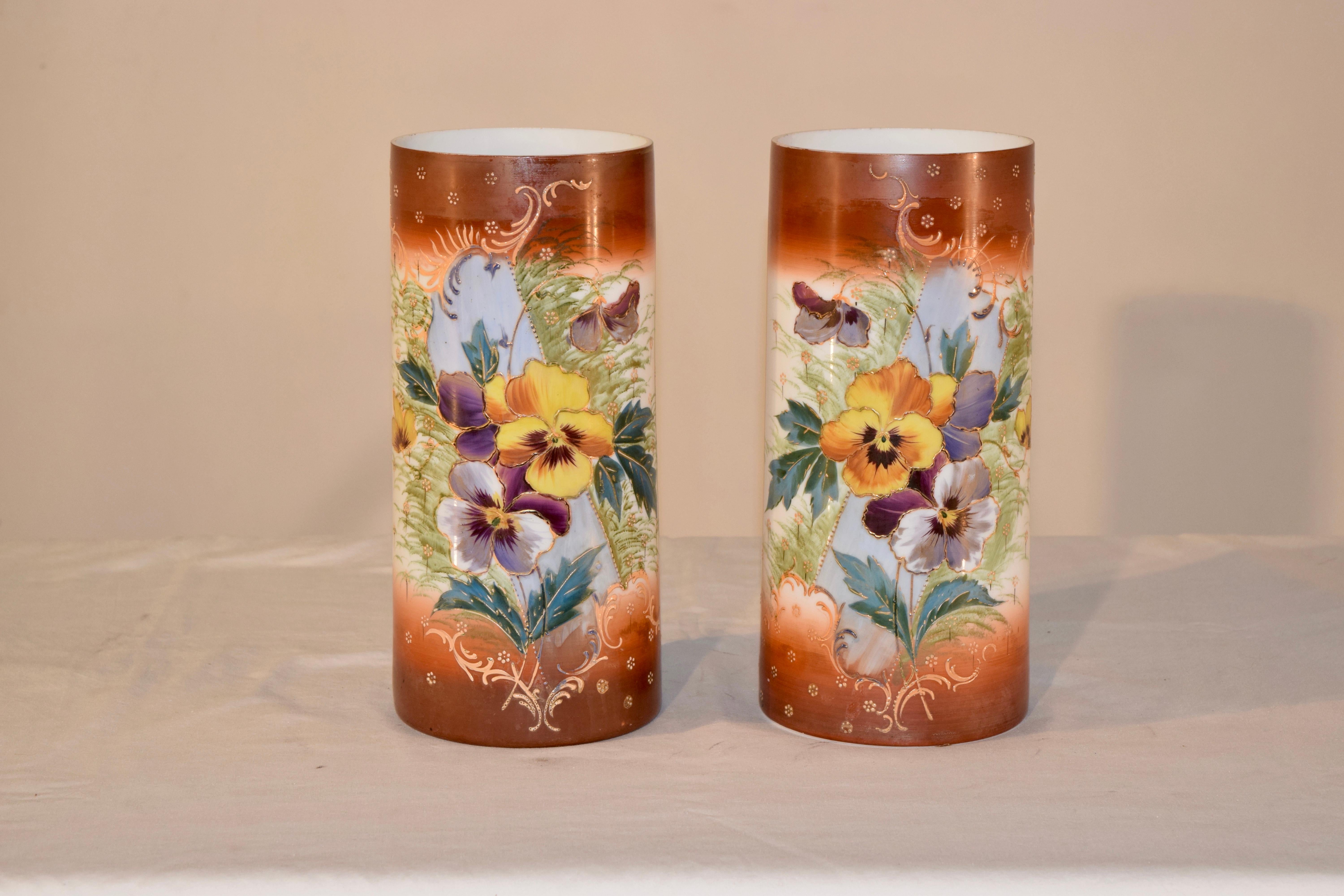 Pair of cylindrical opaline glass vases from France with lovely borders of a warm rust coloring surrounding lovely hand painted pansies and leaves on the front of the vases. There is hand painted gilt accenting as well.