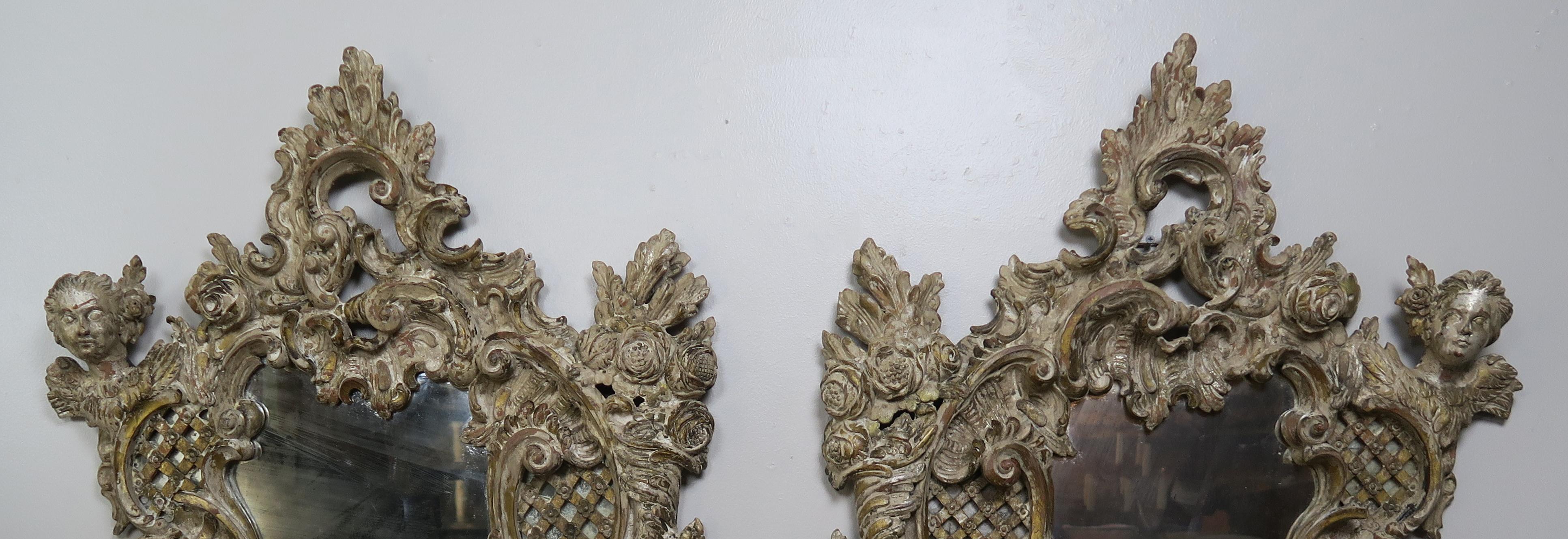 Pair of 19th Century Rococo style Italian Carved Mirrors with Cherubs. The hand carved mirrors are detailed with roses, acanthus leaves, cherub faces and so much more. Beautiful distressed finish with bits of silver leaf and paint remaining.