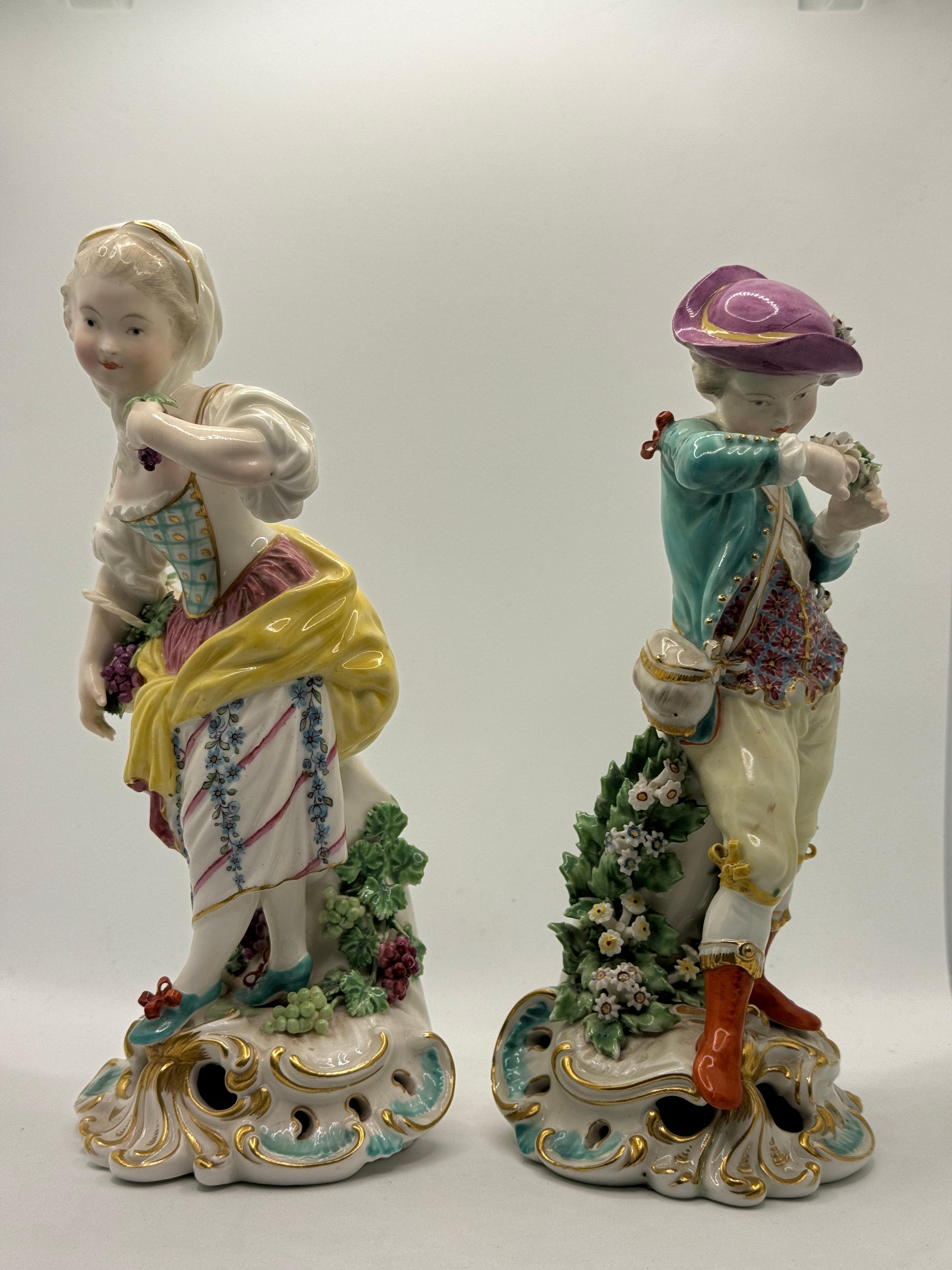 Pair of 19th Century Samson Derby Figures of Autumn & Spring, A girl is depicted as Autumn, in a green bodice and flower decorated skirt with puce sash, with a bunch of grapes in one hand, holding a basket of grapes with the other, beside a bocage