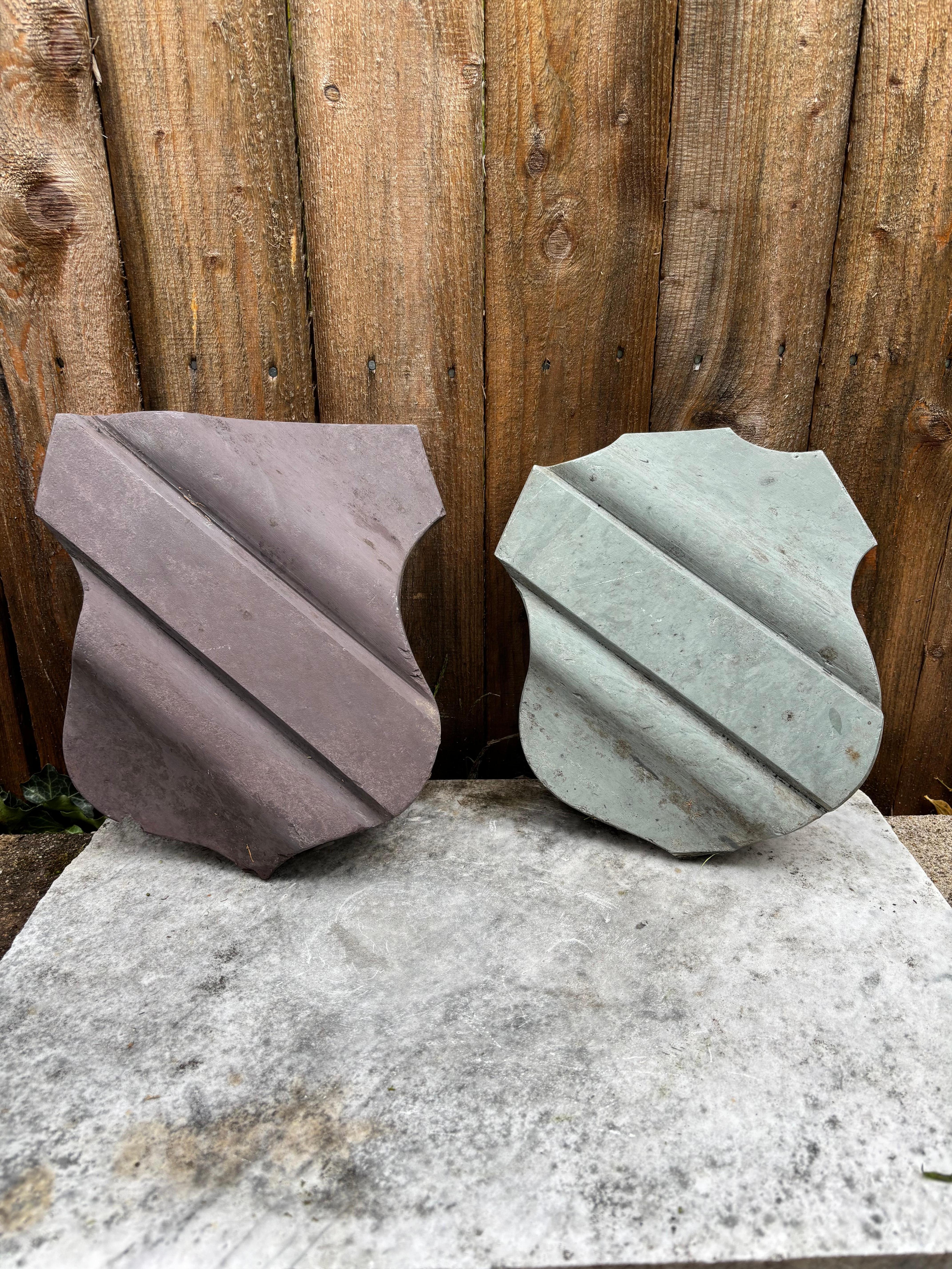 Pair of carved slate architectural elements in the form of armorial shields. Each armorial carved in deep relief with a wide band prominent across the body. Each stylized coat of arms has a distinct shape. One carved from a reddish purple stone, the