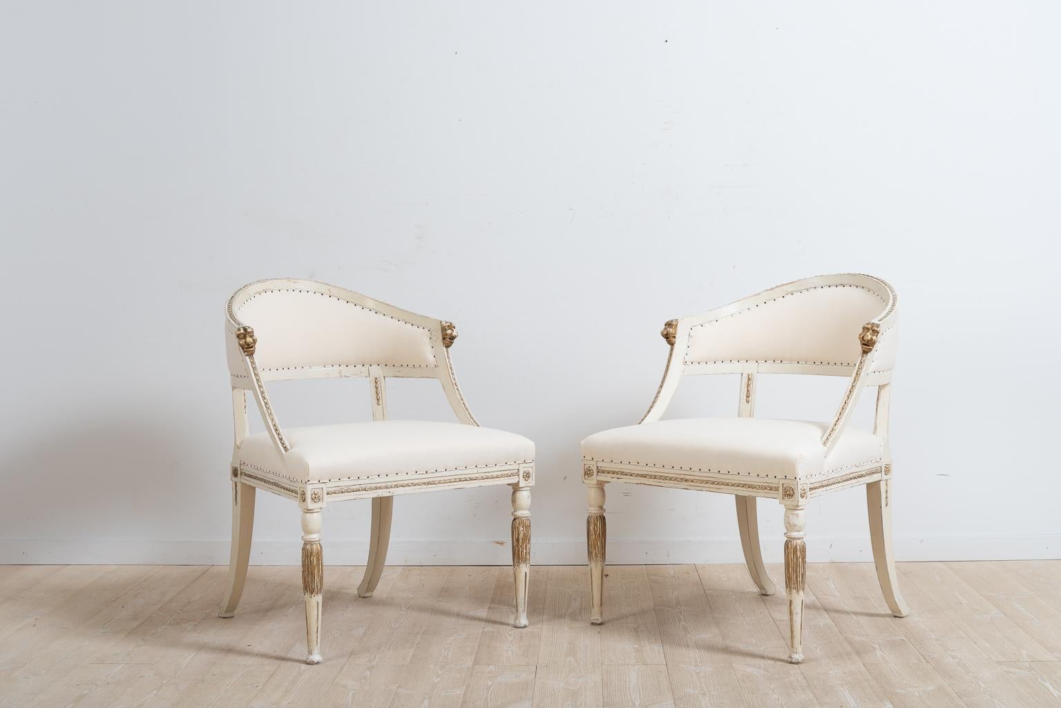 A pair of barrel back armchairs from northern Sweden. The chairs were manufactured in Gustavian style, circa 1880. Carved wooden decorations such as the gold painted lion heads at the end of each armrest. The chairs have their original paint which