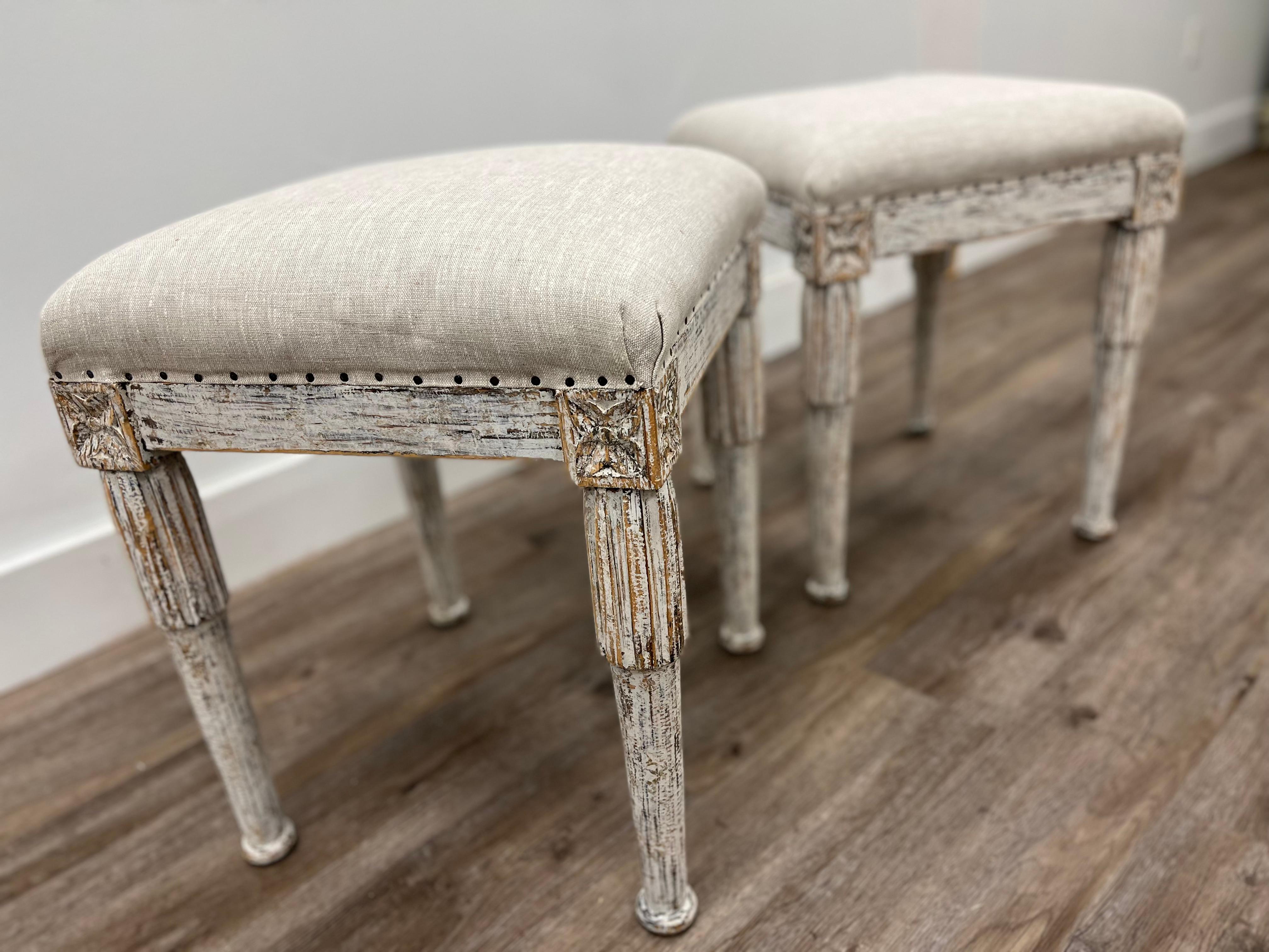 A pair of 19th century Swedish Gustavian style foot stools with detailed fleurons on the corners. The leg tops are finished with vertical reeded over-collaring atop turned and slightly tapered bases. Repainted and newly reupholstered in linen. 
