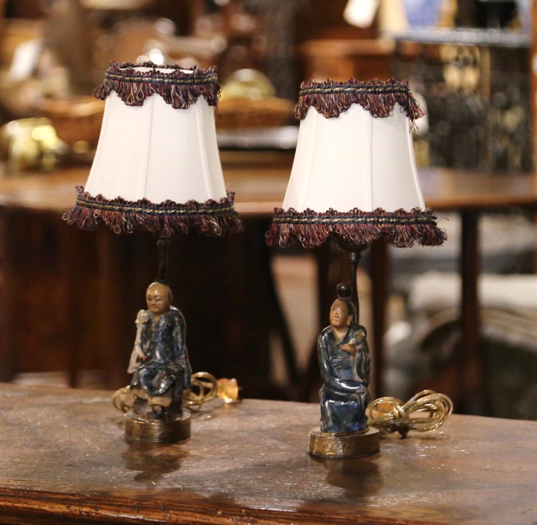 Place these colorful antique lamps on bedside tables, or use them on shelves in a library or study. Created in Asia circa 1890, one lamp stands on a intricate brass base, and depicts an Asian man and woman, both in a sited position. Both petite