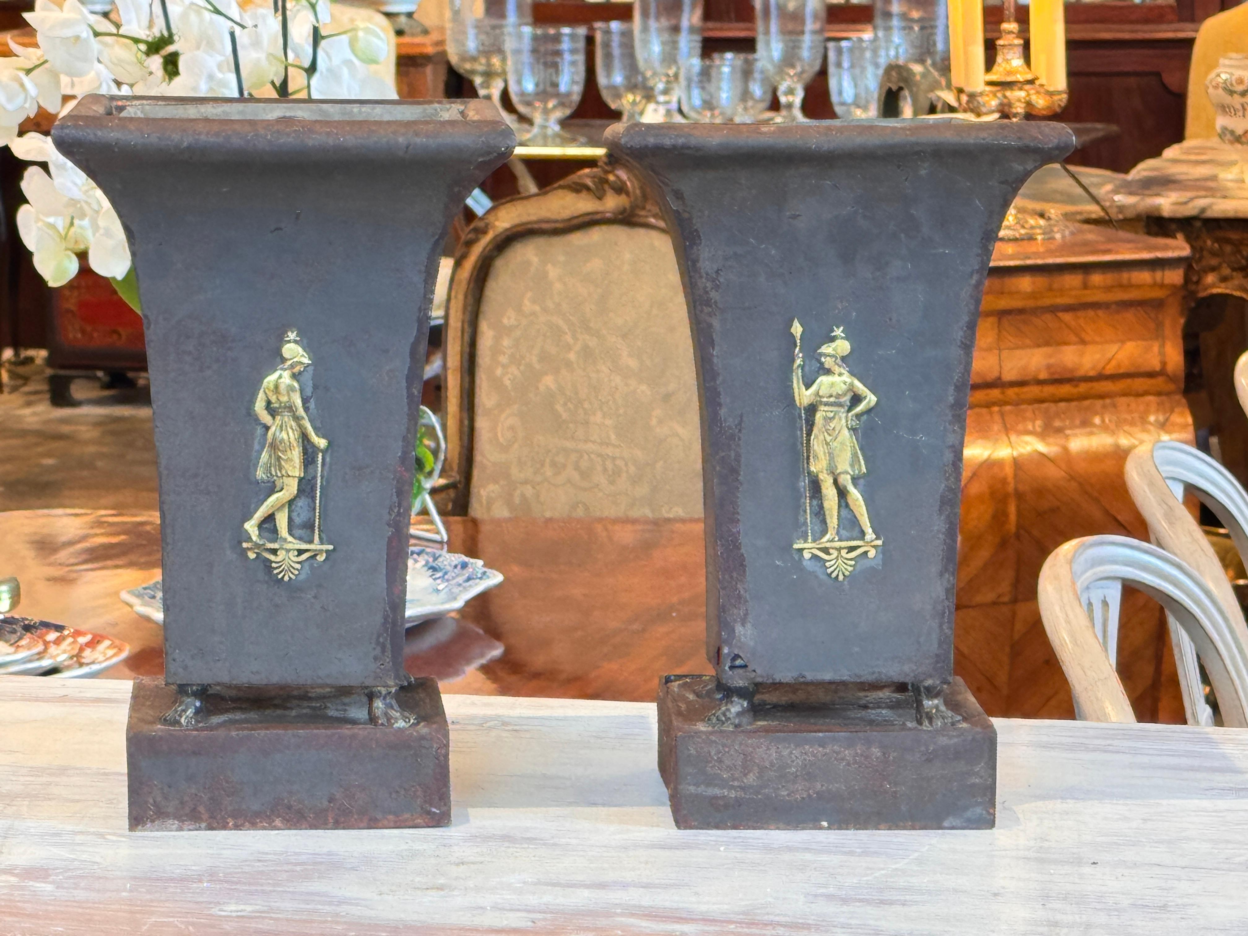 Paire d'urnes en tole brute, chacune décorée de figures classiques dorées en relief. La forme évasée repose sur une base carrée, qui présente une patine vieillie correspondant au travail du métal au XIXe siècle. Un exemple montre la construction