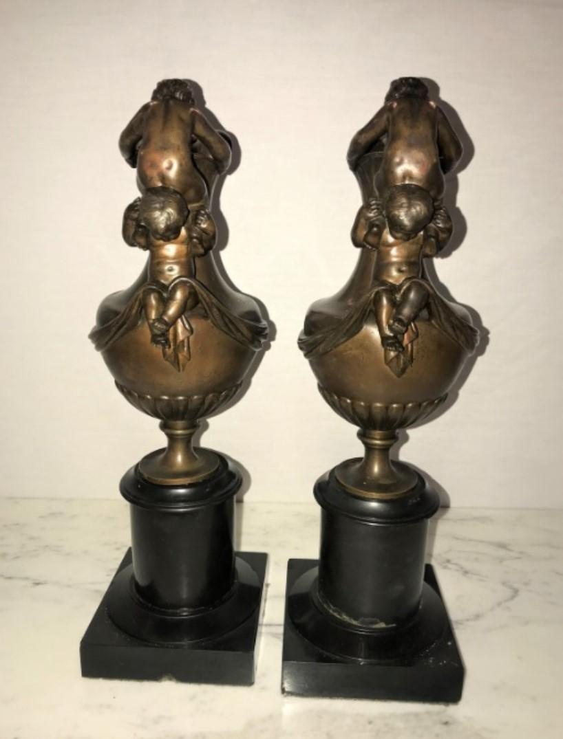 Pair of 19th century French urns on marble stands bearing cherubs and rams heads. Each of these finely chased and bronze patinated urns or water pictures were previously mounted as table lamps and can easily be converted again. The solid black