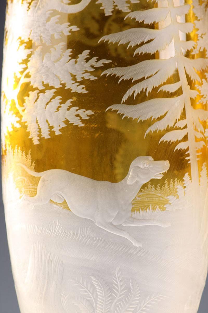 Pair of yellow, Bohemian crystal vases depicting hunting scenes of deer and dogs in the wild. Each vase rests on an elevated base that has undulating edges. The exceptionally detailed work highlights the patterns engraved in the transparent crystal.