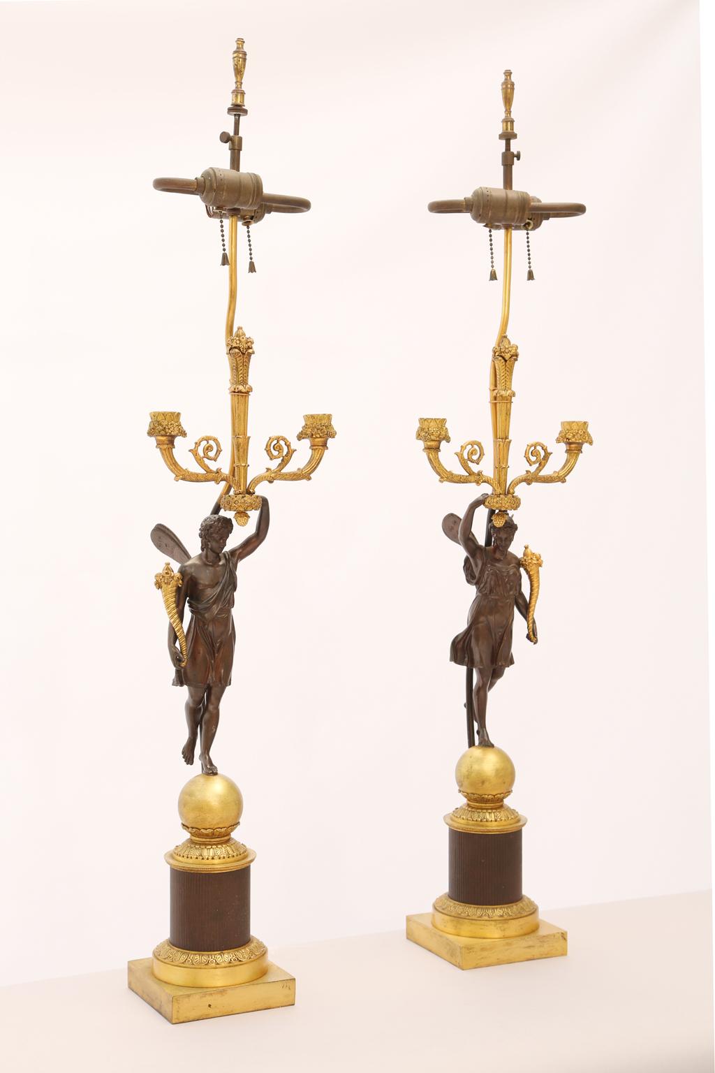 Opposing pair of electrified figural candelabras in gilt and patinated bronze, each feature a winged, patinated bronze nymph, standing upon a sphere, holding a cornucopia and bouquet, surmounted by double candle branches, raised on foliate
