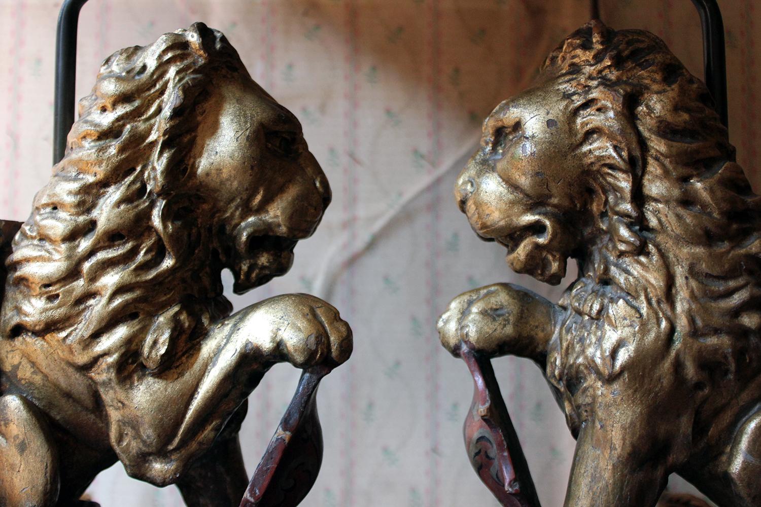 The fine pair of 19th century French carved softwood gilded lions, each in the passant position with their right for paw raised over painted scarlet armorial shaped crests showing a variant of the fleur-de-lys, probably having once been part of an