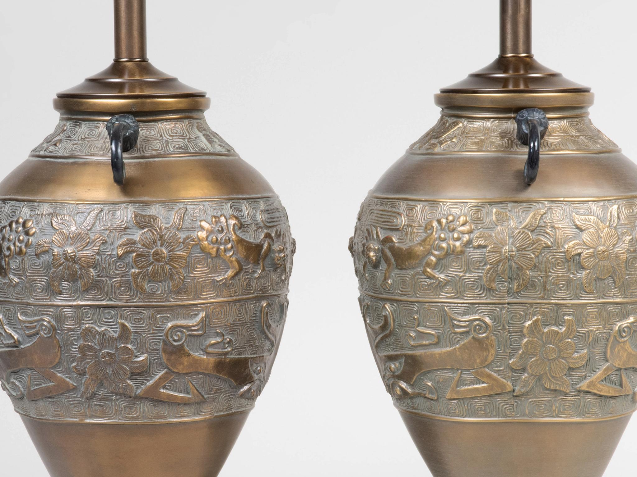 An ornate 20th century pair of table lamps in the manner of James Mont. They are comprised of cast brass bodies, ebonized wood bases, and new custom linen shades. Stylized floral and animal figures adorn the body of the lamps, while the double