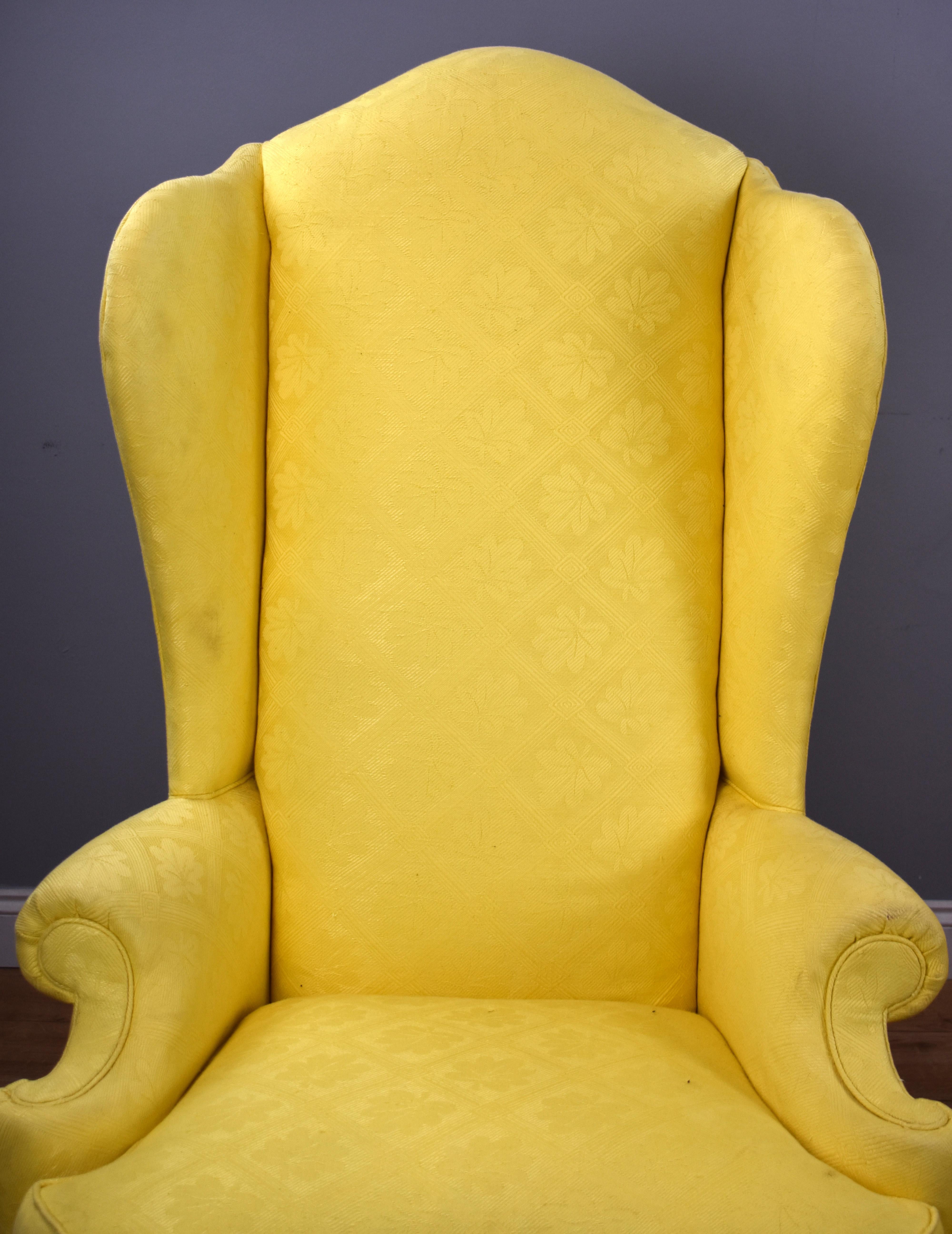 A pair of Queen Anne style wing chairs, opholstered in a yellow fabric and standing on elegant cabriole legs united by a stretcher with a carved finial in the centre. Both chairs are structurally sound and in good order, showing minor signs of wear