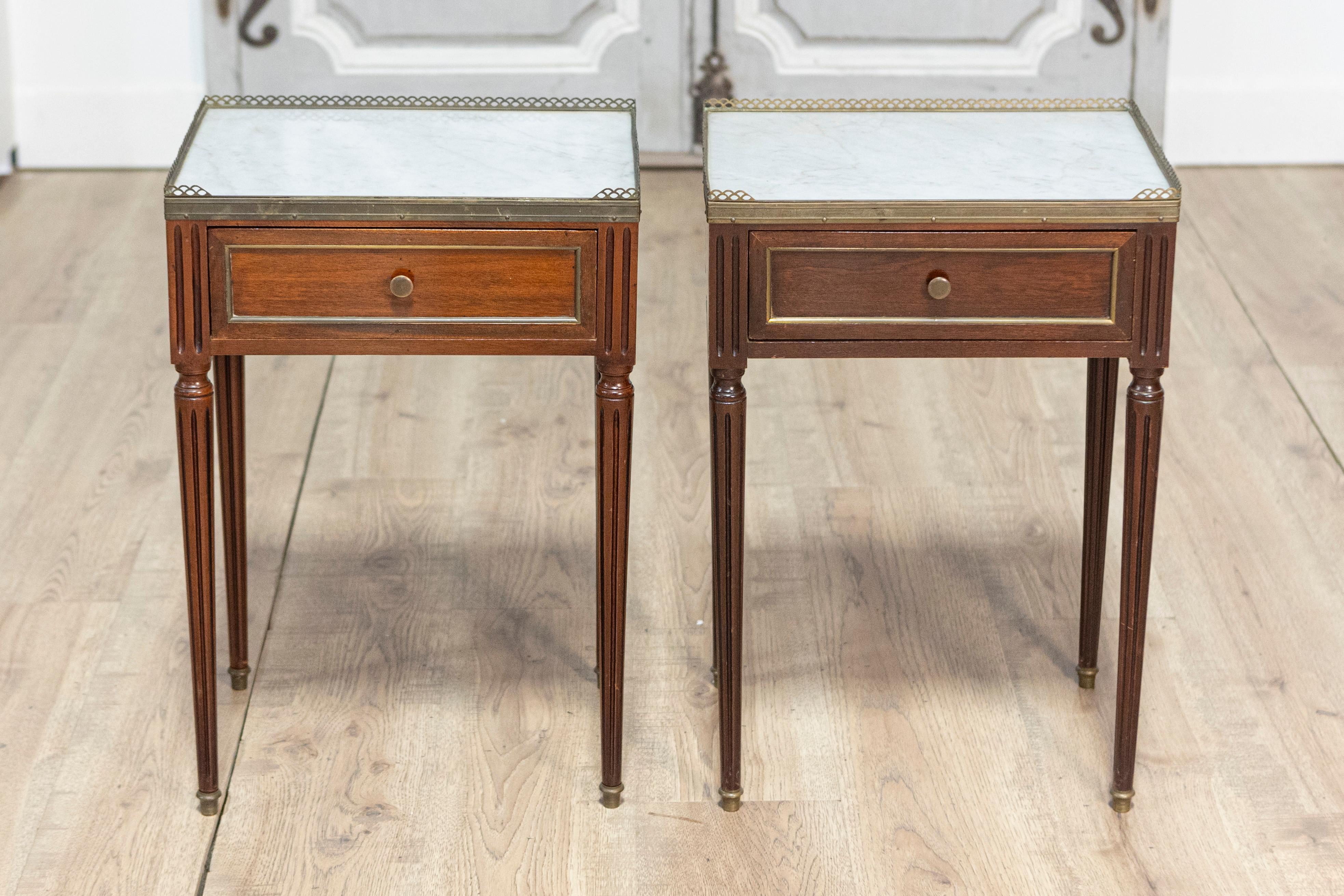 This refined pair of 20th century French bedside tables embodies the timeless elegance of the Louis XVI style. Topped with pristine white marble and encircled by a delicate brass gallery rail, they strike the perfect balance between sophistication