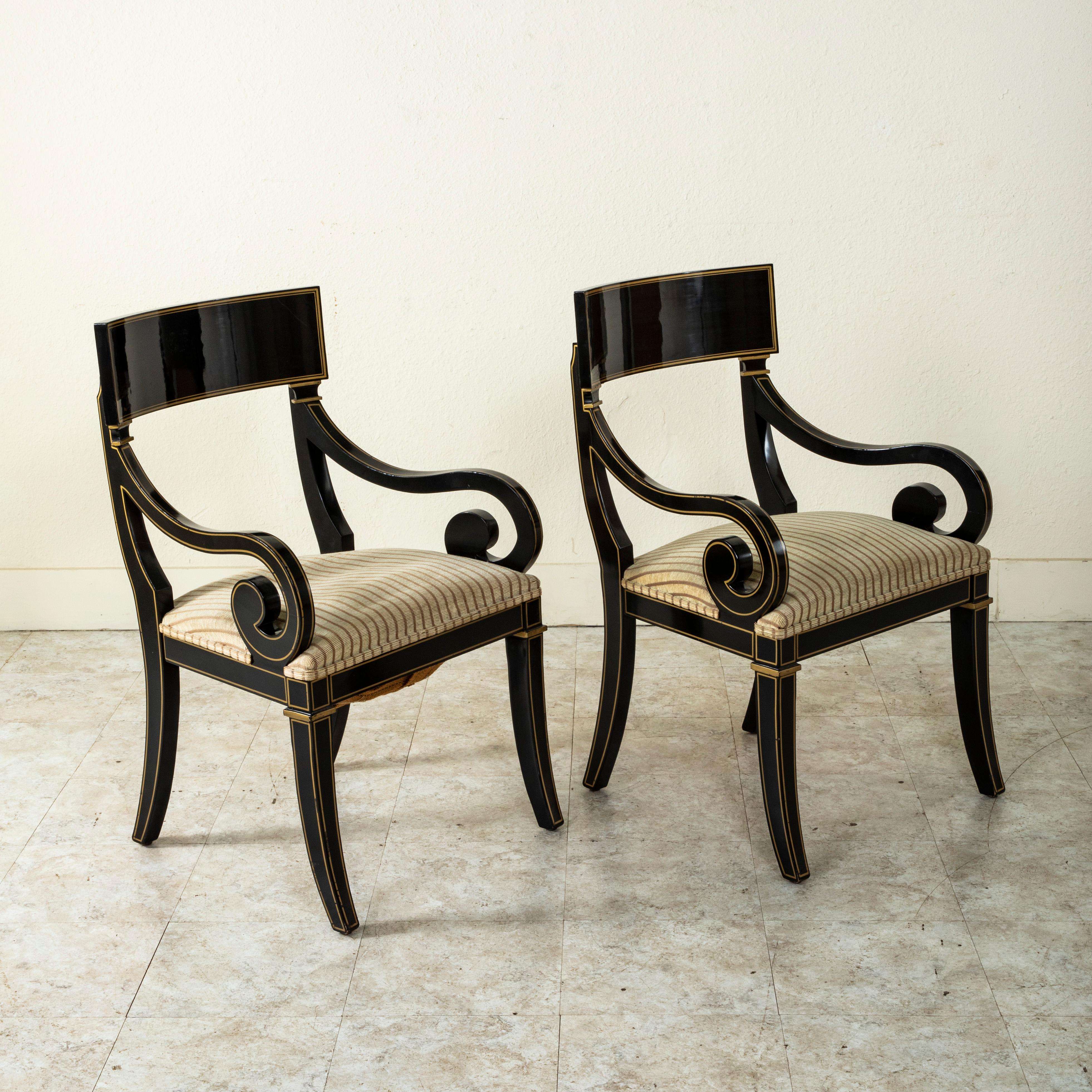 This pair of mid-twentieth century French Restauration style armchairs features a black lacquered finish with gold detailing. The pair is detailed with square capitals below the seat backs, scrolling armrests and legs that gently curve outward 