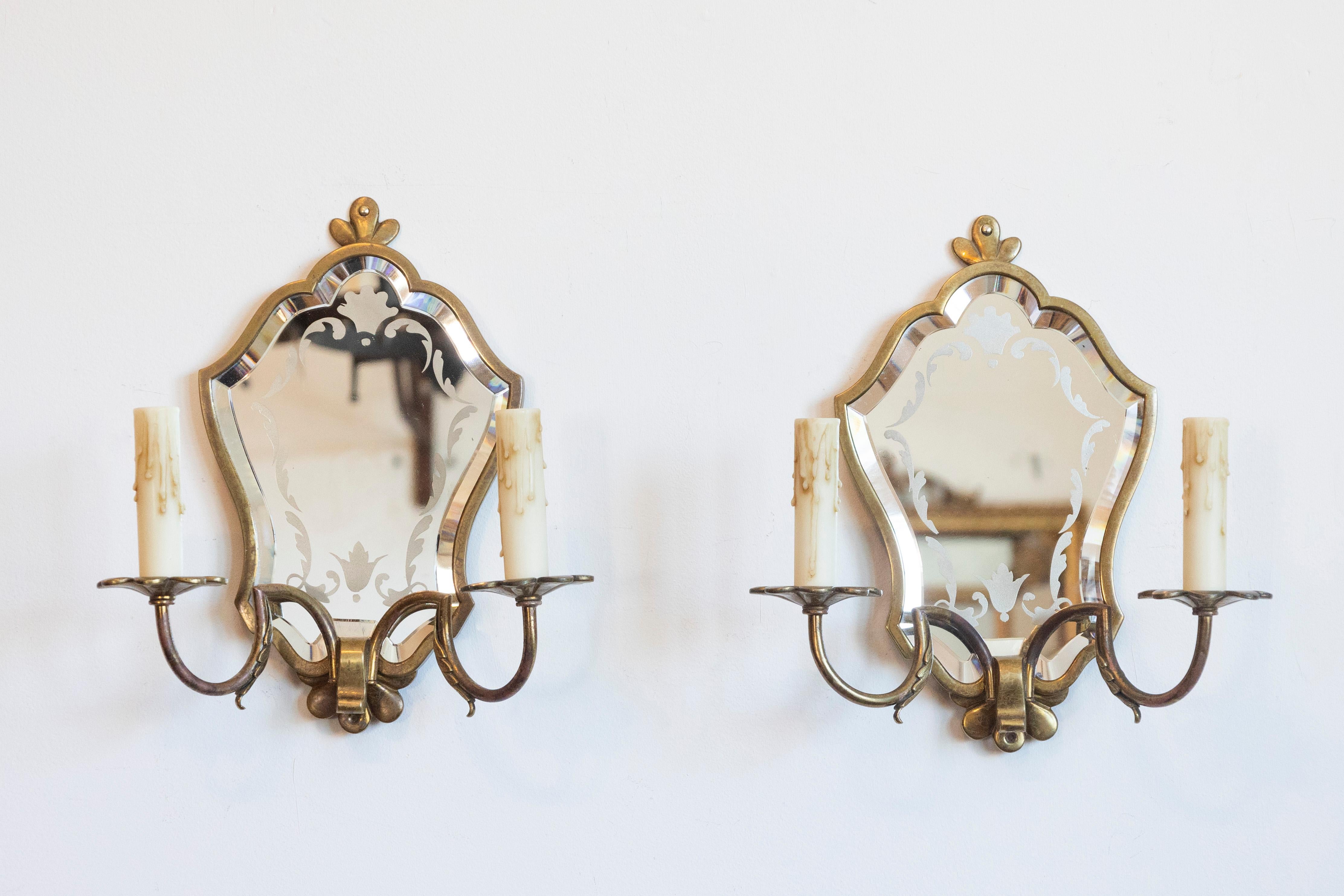 Cette élégante paire d'appliques miroir italiennes en laiton du XXe siècle incarne l'artisanat européen classique avec une esthétique raffinée et décorative. Chaque applique présente un miroir biseauté de forme ornementale avec des détails gravés,