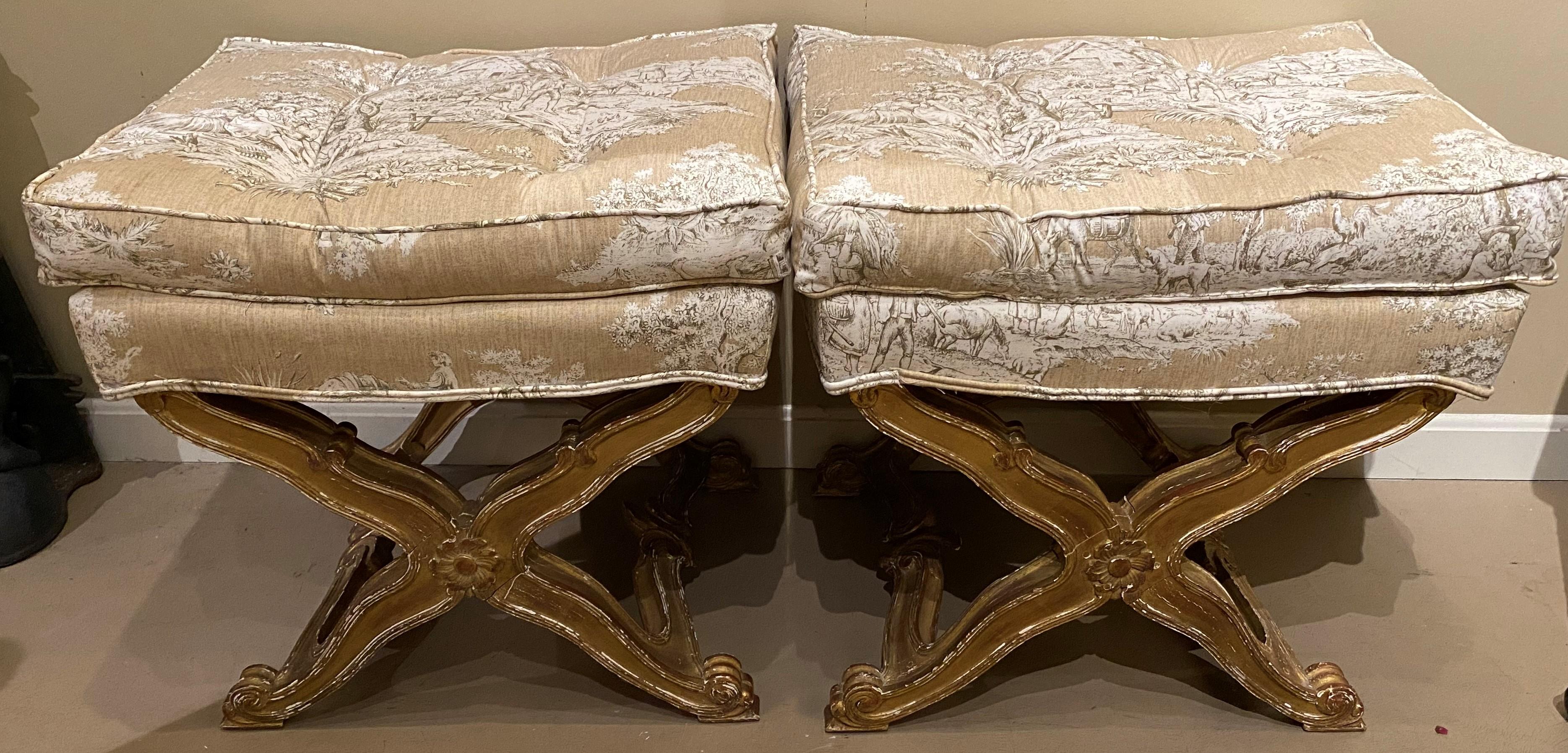 An attractive pair of Italian curule style Ottomans with giltwood bases and decorative recent tufted upholstery with genre scenes. Very good overall condition, with some gilt losses on the bases, high point edge losses, and light wear commensurate