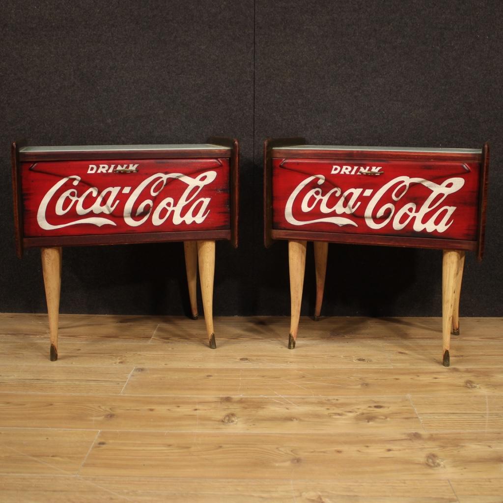 Pair of Italian design nightstands from the 1960s-1970s. Furniture with walnut sides, light wood legs, colored glass tops and front door painted with the Coca Cola logo (not original), recently painted logo. Fall-front door that offers a good