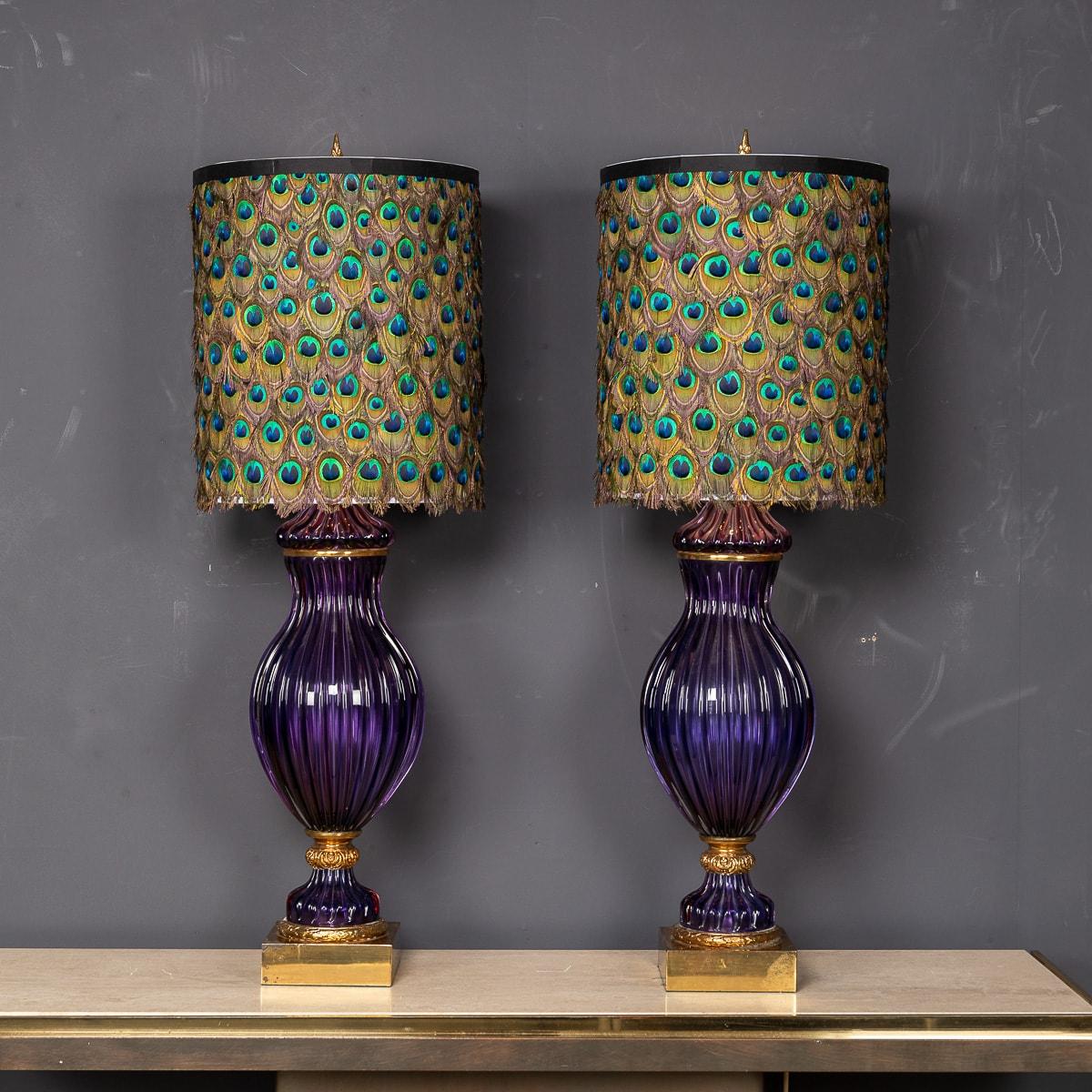 A wonderful mid 20th Century pair of Murano glass neoclassical lamps. The glass, a handblown purple urn shape, gilt with details have a double bulb fitting with original chain switches. The shades have been updated to an opulent peacock feather