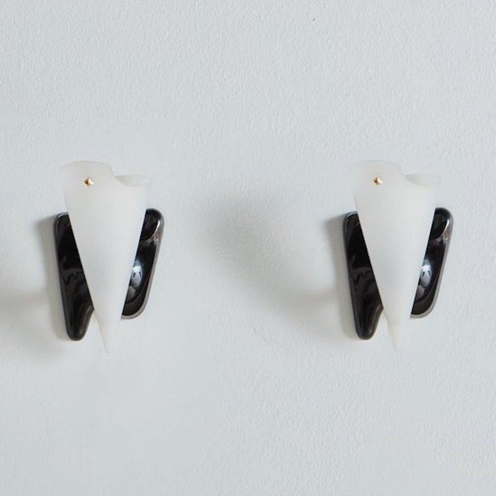 A pair of 1950s French wall sconces in the style of Pierre Guariche. These sconces feature curved black acrylic bases with adjustable ribbed white acrylic cone shades. Each shade has a subtle brass decorative detail and brass hardware.
