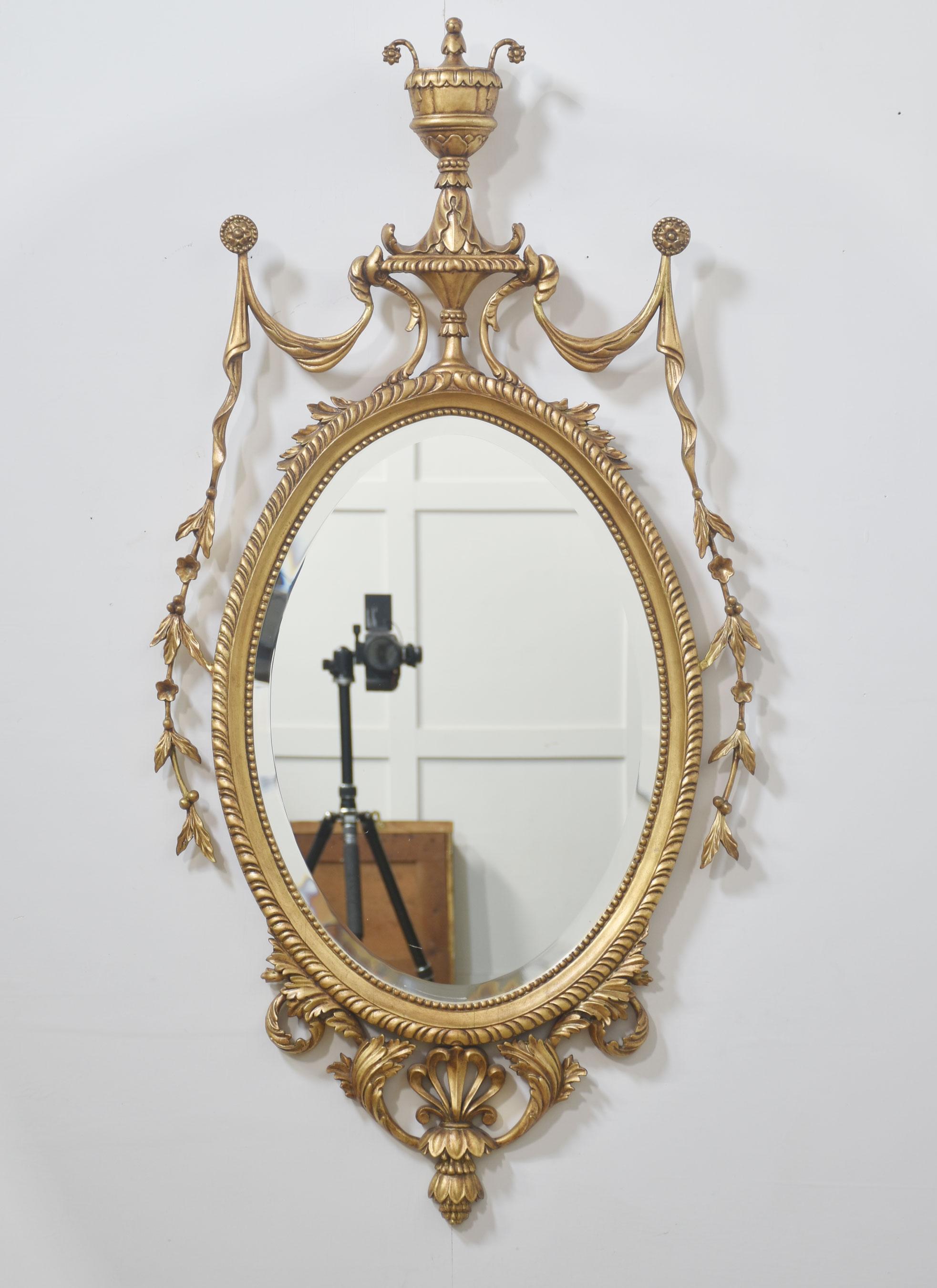 Pareja de espejos de pared ovalados de madera dorada tallada estilo Adam. Cada espejo tiene una placa ovalada de cristal biselado dentro de un marco estriado, coronado por un remate clásico en forma de urna y adornado con espirales de cintas, gotas