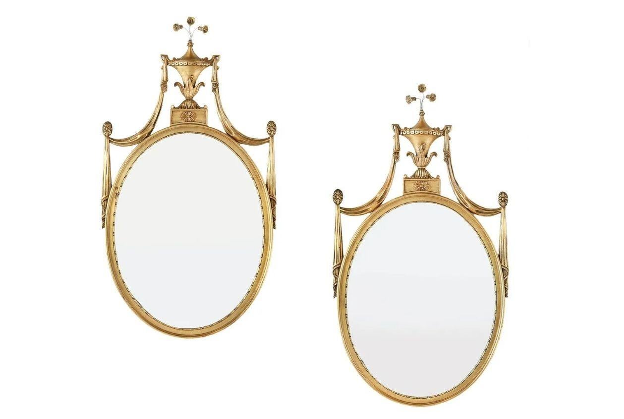 Pair of Adams style giltwood wall mirrors, console or commode mirrors
 
Each mirror having an oval frame flaked by swag carving terminating in an urn and scroll floral design. A classic Adam style crest with acanthus leaf and palmette on top of