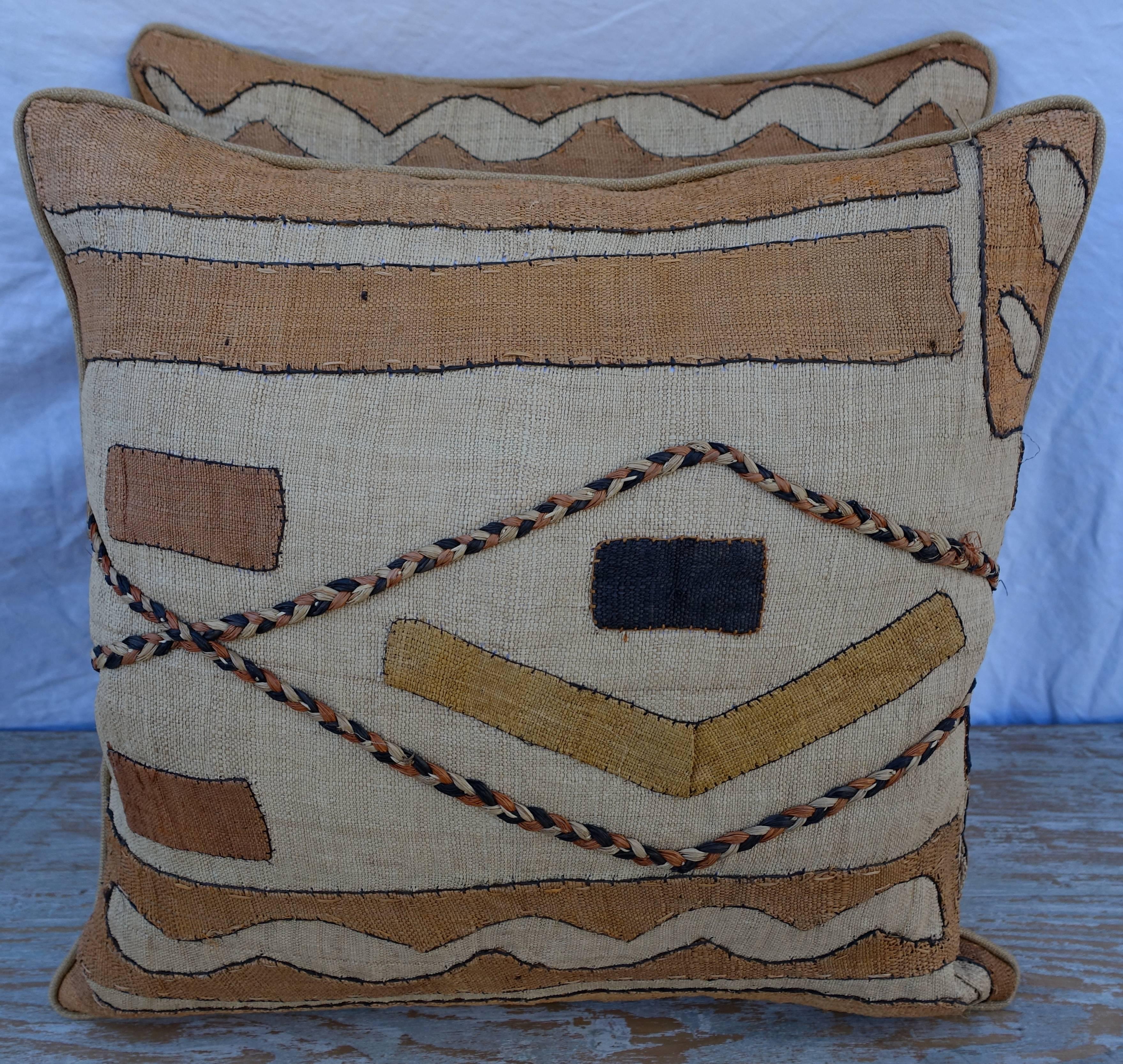 Pair of African Kuba cloth pillows with raffia Kuba cloth fronts in black, gold and cream coloration and golden linen backs. The pillows are finished with a self cord detail. Down inserts, sewn closed.