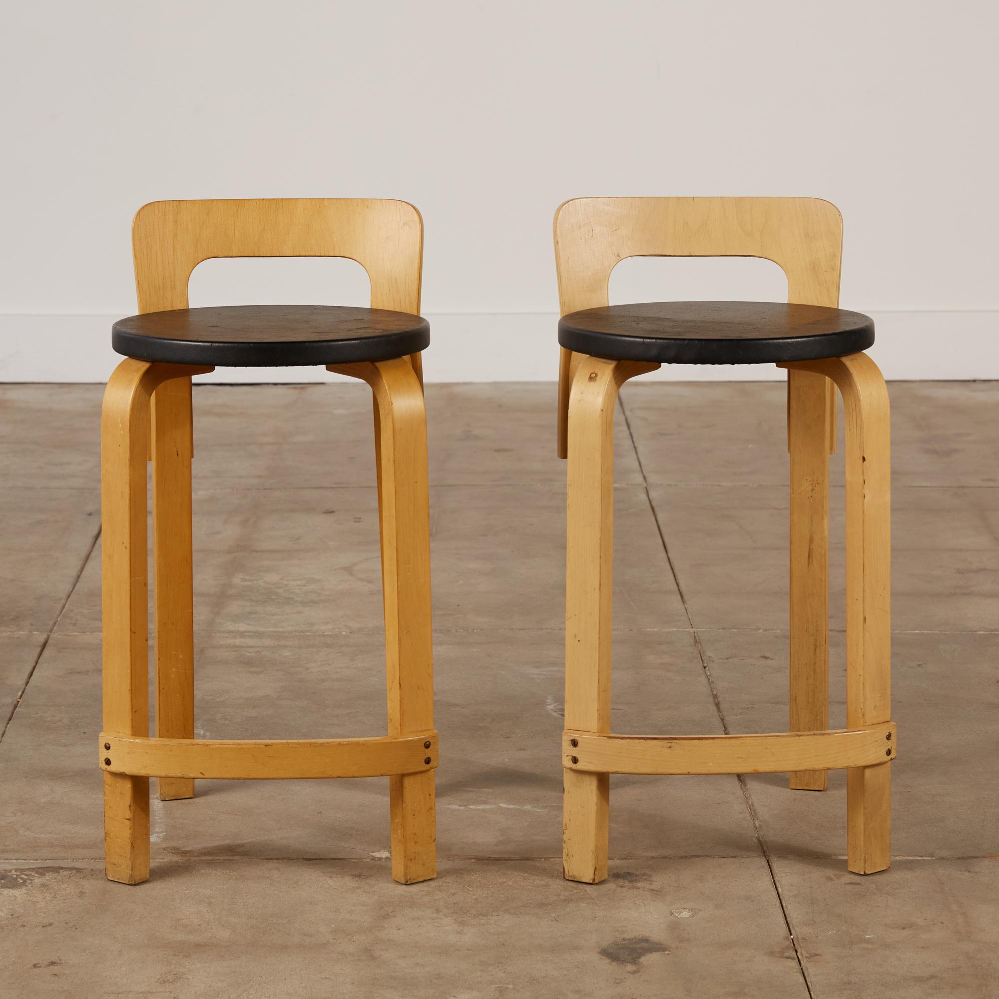 This pair of Artek’s K65 high chairs/stools designed by Alvar Aalto in 1935 and produced in the 1950's, Sweden, feature honey toned birch legs and a low bent plywood backrest. The seat is covered in a patinated black vinyl and has curved legs as