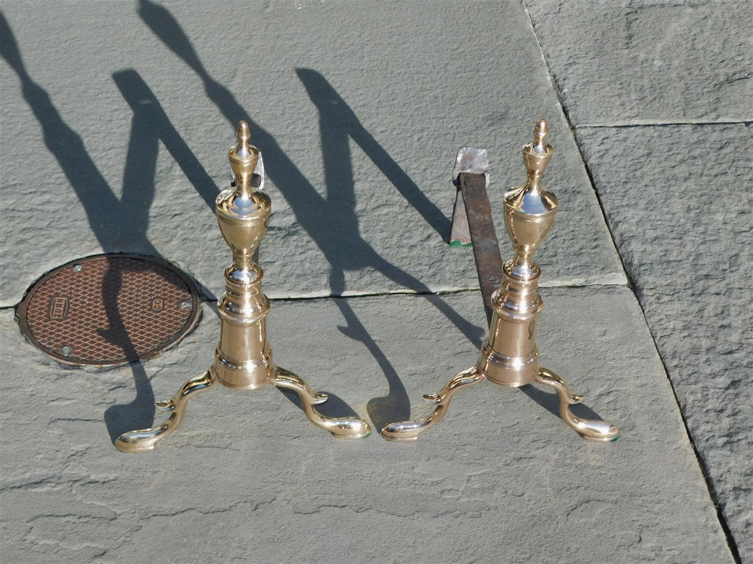 Pair of American bell brass double urn finial andirons with circular ringed plinths, spur legs, wrought iron rear dog legs, and resting on original slipper feet. New York, Late 18th Century. Wrought iron dogs legs can be adjusted in length by