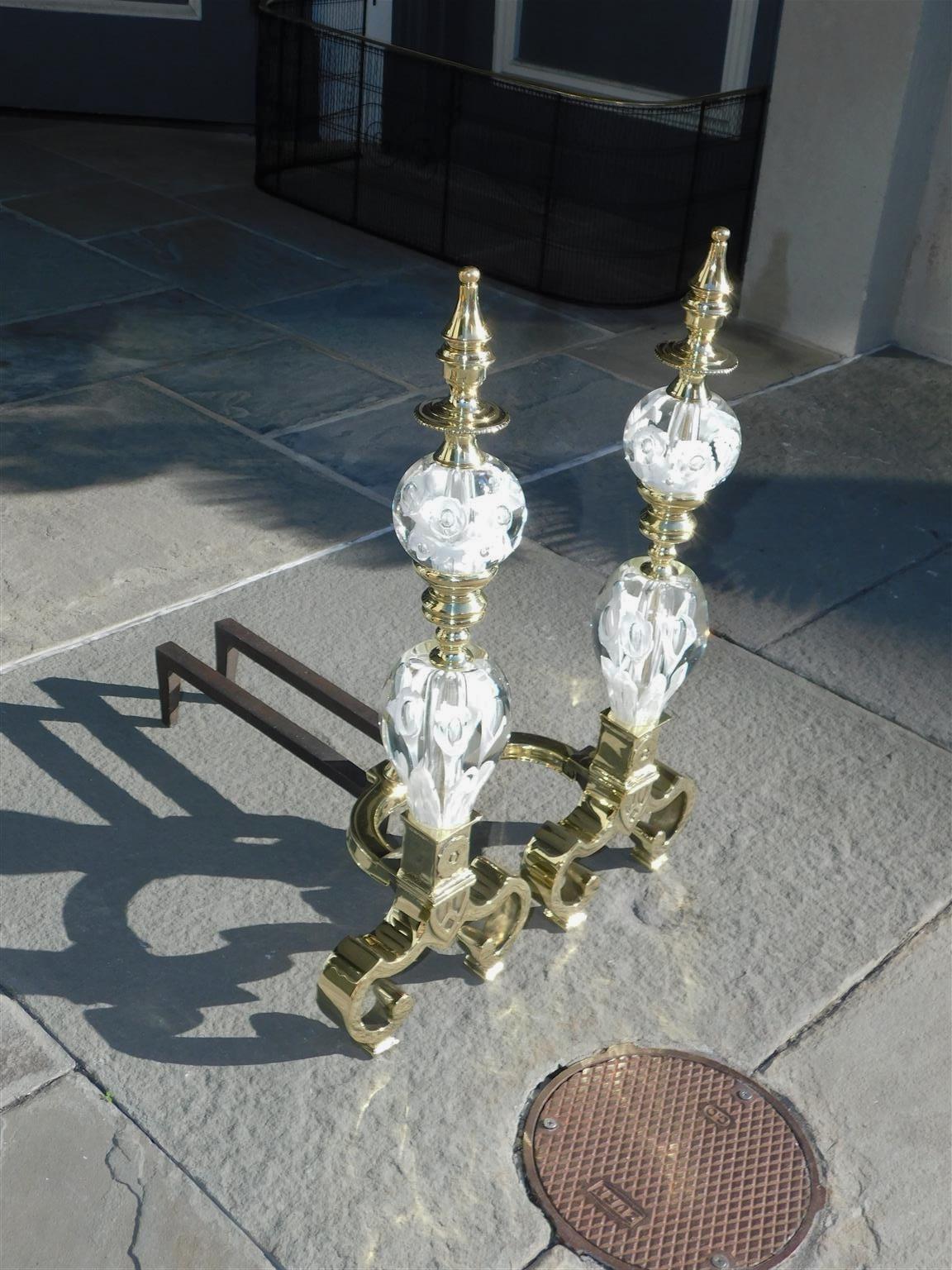 Pair of American brass St. Clair andirons with clear globes containing controlled interior bubbles featuring groupings of stylized white flowers. Andirons consist of flanking beaded urn finials, crystal sphere balls, turned bulbous columns, and