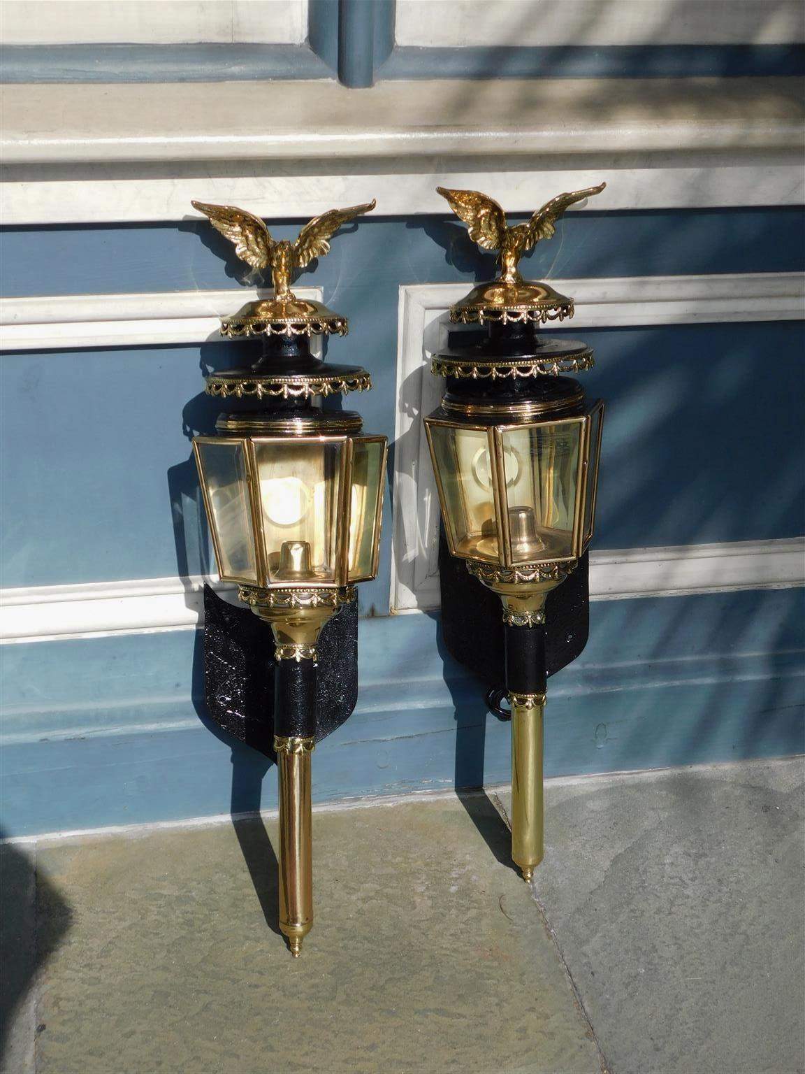 Pair of American brass coach lanterns with flanking eagle finials, decorative tiered beaded swags, original beveled glass, and mounted on cuffed shield back scrolled iron brackets. Mid 19th Century
Pair of lanterns have been lacquered for exterior