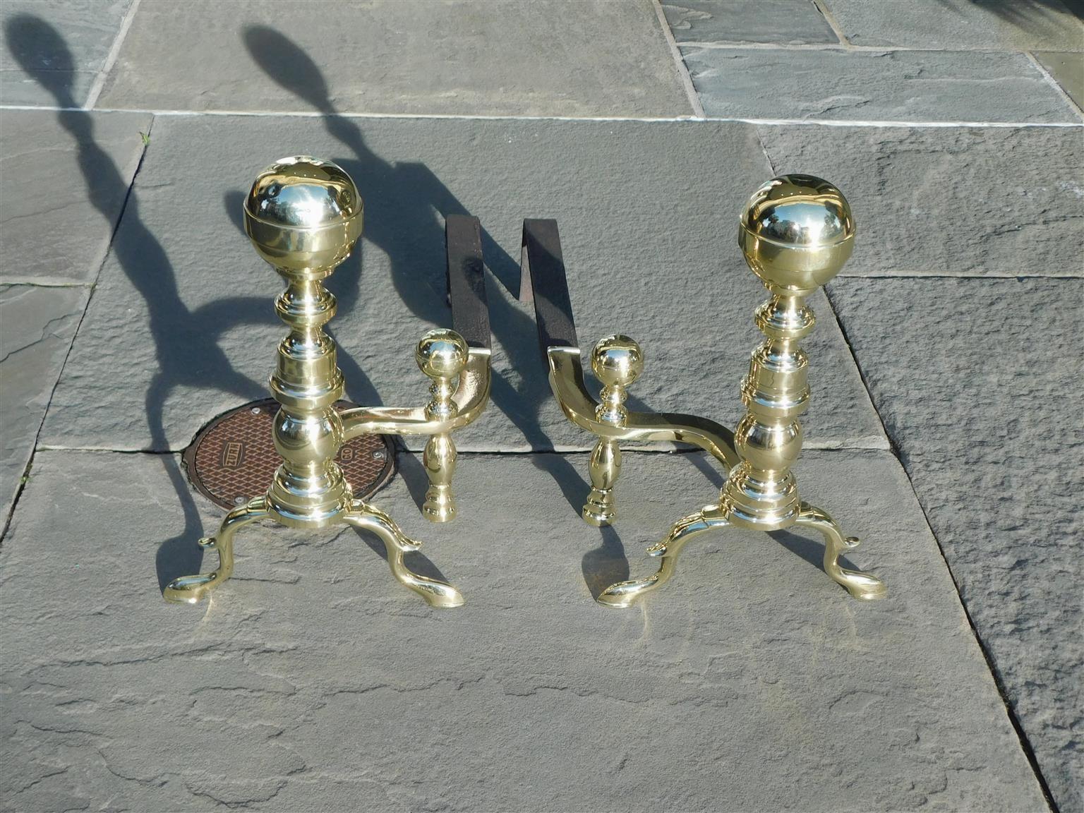 Pair of American brass ball finial andirons with bulbous ringed plinths, spur legs, curvature matching log stops, wrought iron dog legs, and resting on original slipper feet. Boston, MA. Early 19th Century. Wrought iron dogs legs can be adjusted in