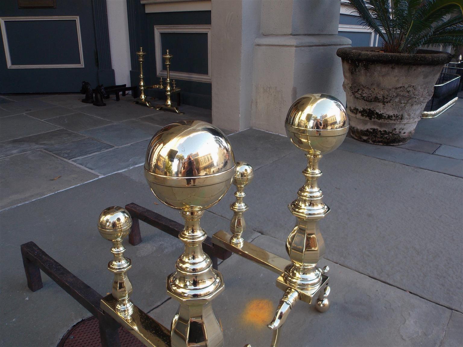 Pair of American brass ball banded finial andirons with turned bulbous faceted plinths, matching ball finial faceted log stops, scalloped skirt, original rear iron dog legs, and terminating on banded spur legs with ball feet. Boston, MA, Early 19th