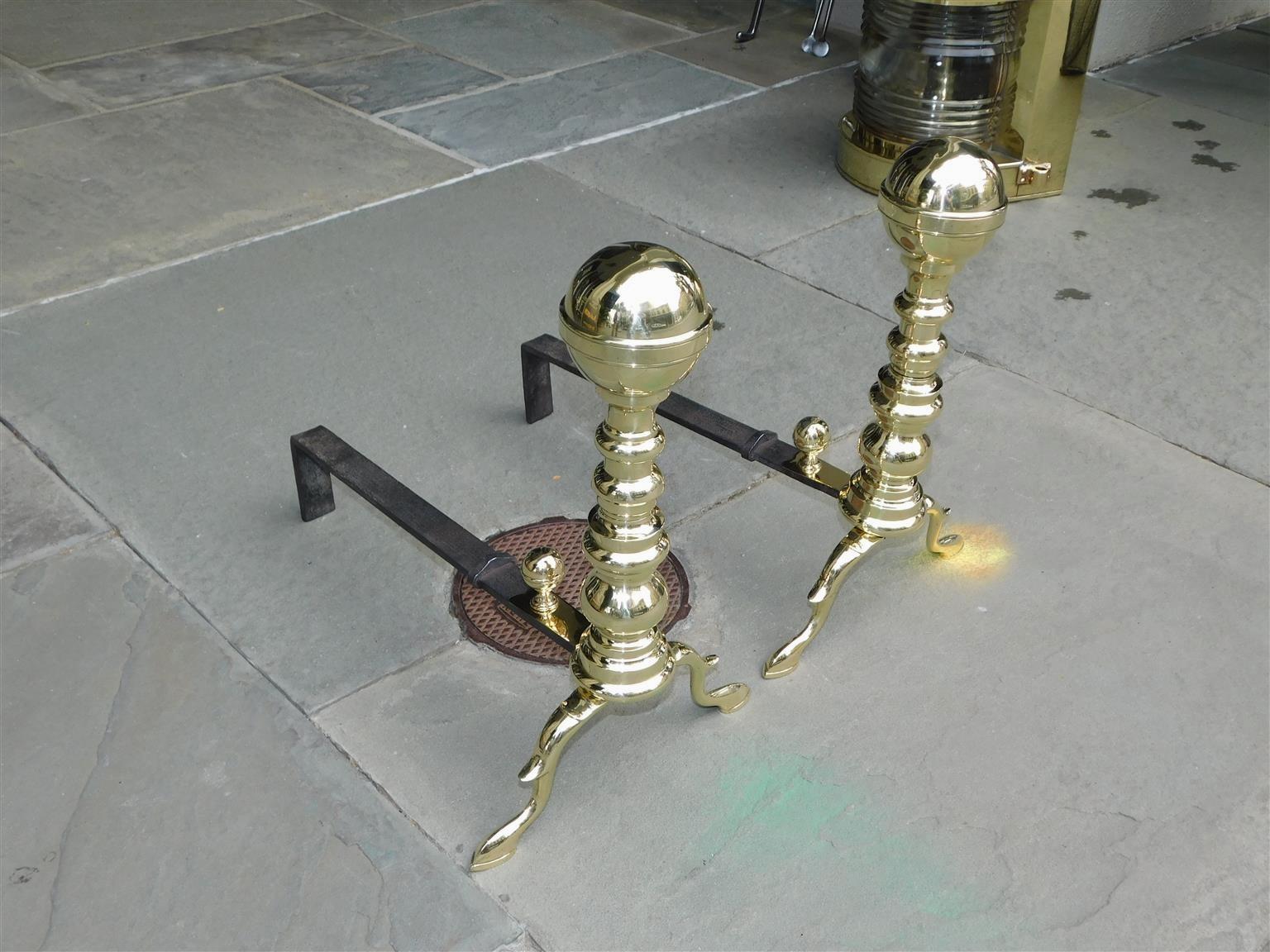 Pair of American brass banded flanking ball finial andirons with turned bulbous plinths, matching ball banded log stops, original wrought iron rear dog legs, and resting on spur legs with slipper feet. Boston, MA. Early 19th Century.