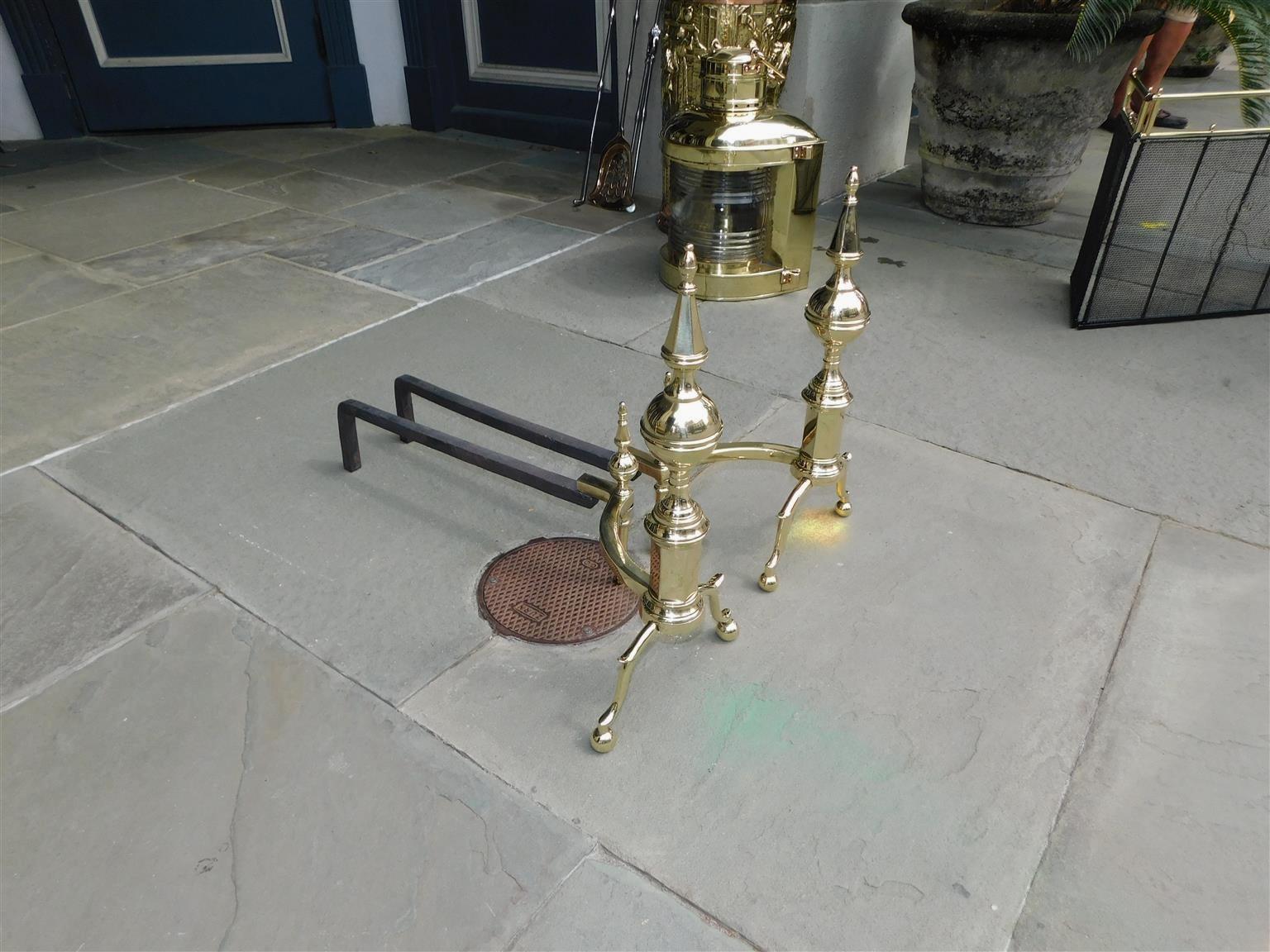 Pair of American brass banded flanking steeple and ball finial andirons with ringed faceted plinths, curvature supporting matching steeple log stops, original wrought iron rear dog legs, and resting on spur legs with ball feet. New York, Early 19th
