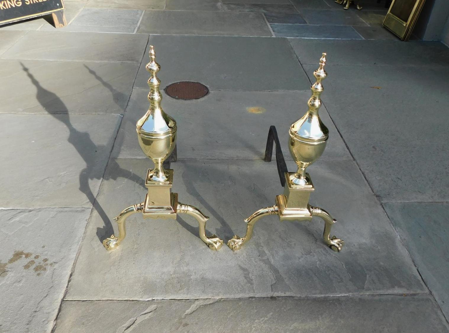 Pair of American brass flanking faceted Urn on Urn finial andirons with squared plinths, scalloped skirts, original wrought iron rear dog legs, and resting on banded spur legs with ball and claw feet. Philadelphia, late 18th century.