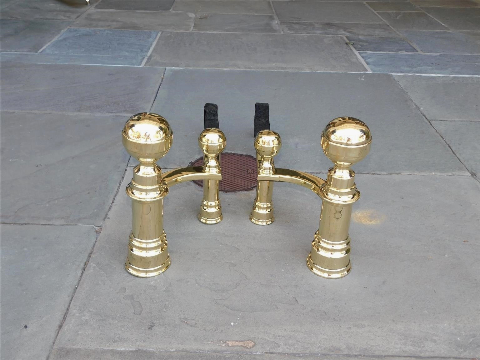 Pair of American brass banded flanking ball finial andirons with circular ringed plinths, matching curvature banded log stops, and resting on the original wrought iron rear dog legs. Stamped by maker John Hunneman, Boston. Early 19th century.