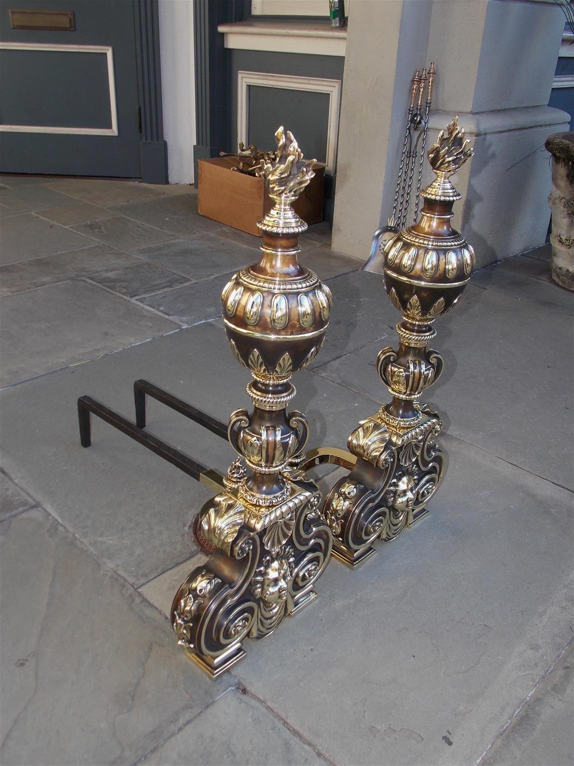 Pair of American bronze and brass ball top andirons with hand chased beading, bossing, foliage, and flanking flame finials. The andirons have a stylized centered scrolled foliage urn resting on a squared gadrooned plinth, matching flame finial log