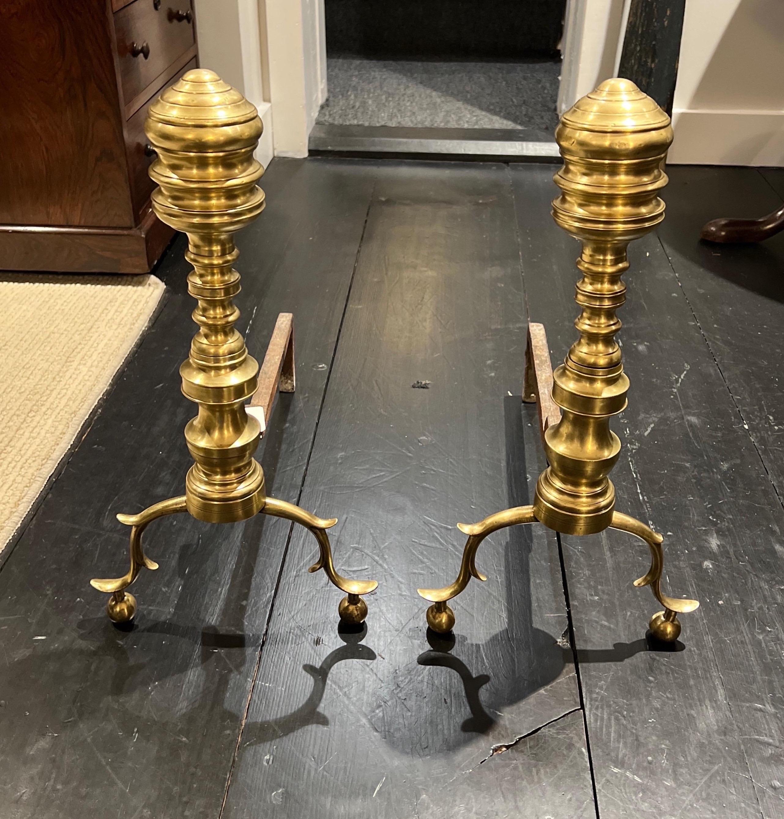 A Pair of American Federal Period finial form brass andirons, both showing turned finial capitals on sectional columns rising from double spurred and hipped supports, both retaining their original forged iron 'dogs',

The brass having a fine patina,