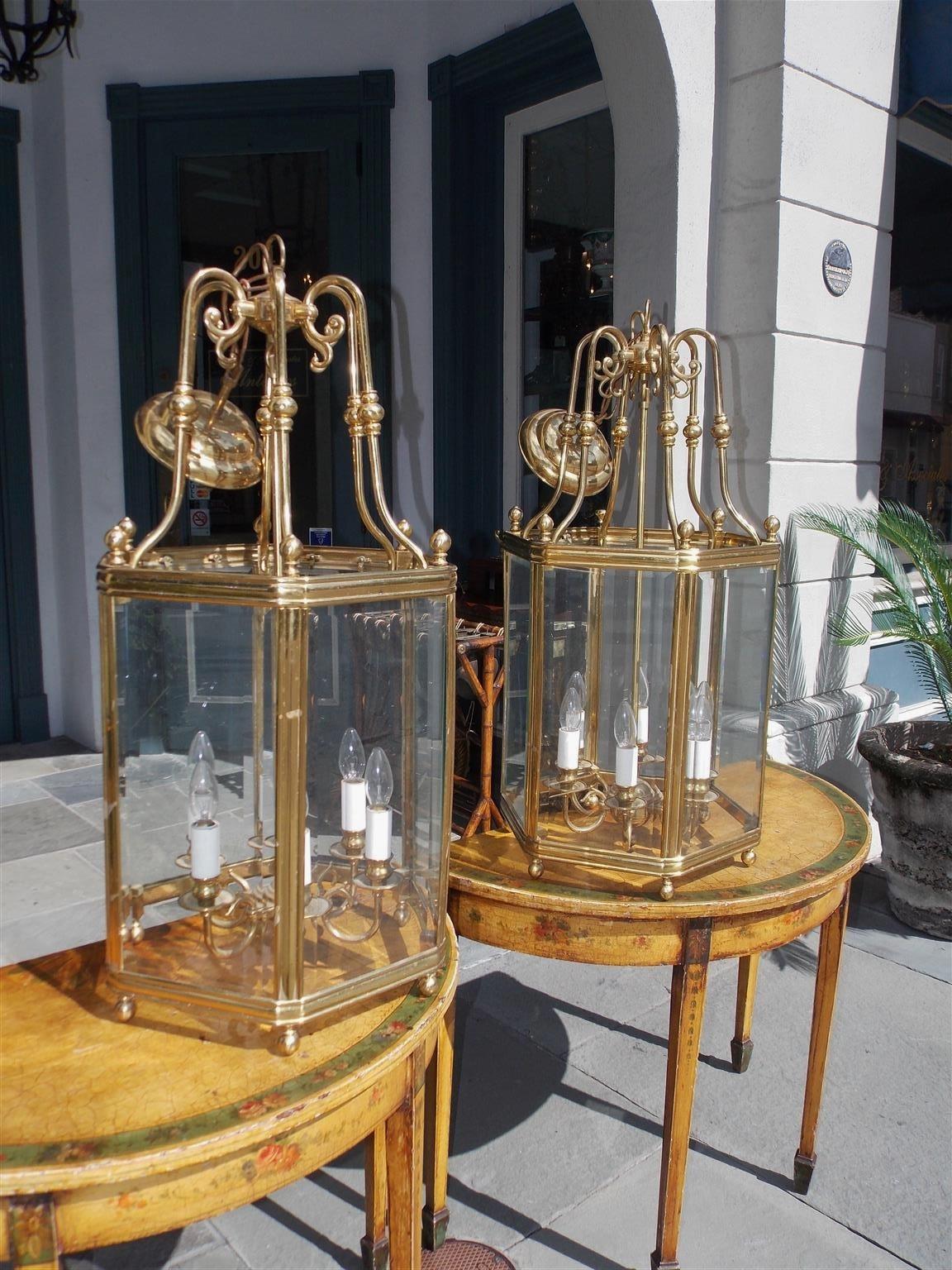 Pair of American brass colonial style hexagon hanging hall lanterns with decorative scrolled bulbous arms, stylized acorn finials, original beveled glass, six light interior cluster, and resting on a molded edge base with ball feet, Late 19th
