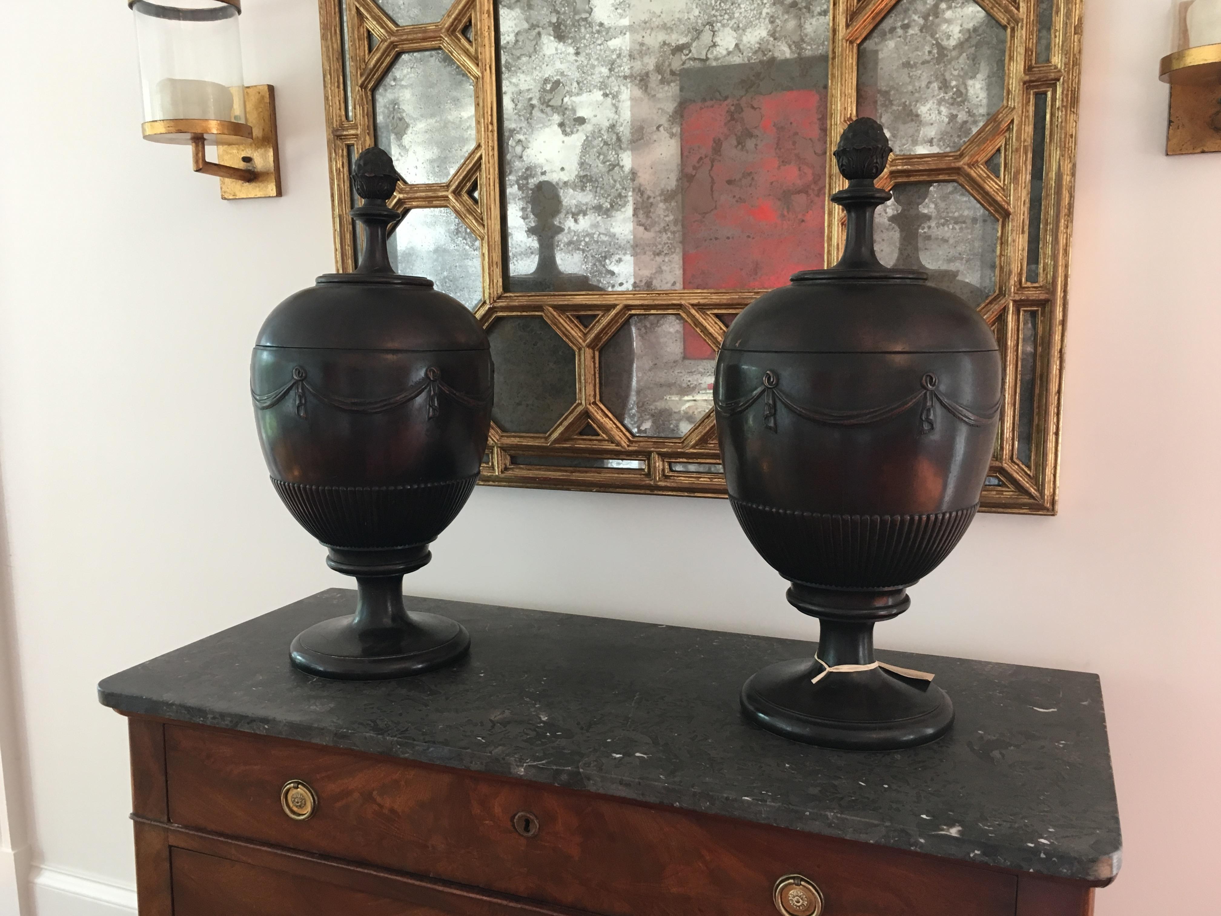Pair of American mahogany urn shaped cutlery boxes with pineapple finials, carved ribbon swags followed by a line of beading, original cutlery inserts, and resting on turned round bases.

One urn has loss at the edge of the top - see photo. Also