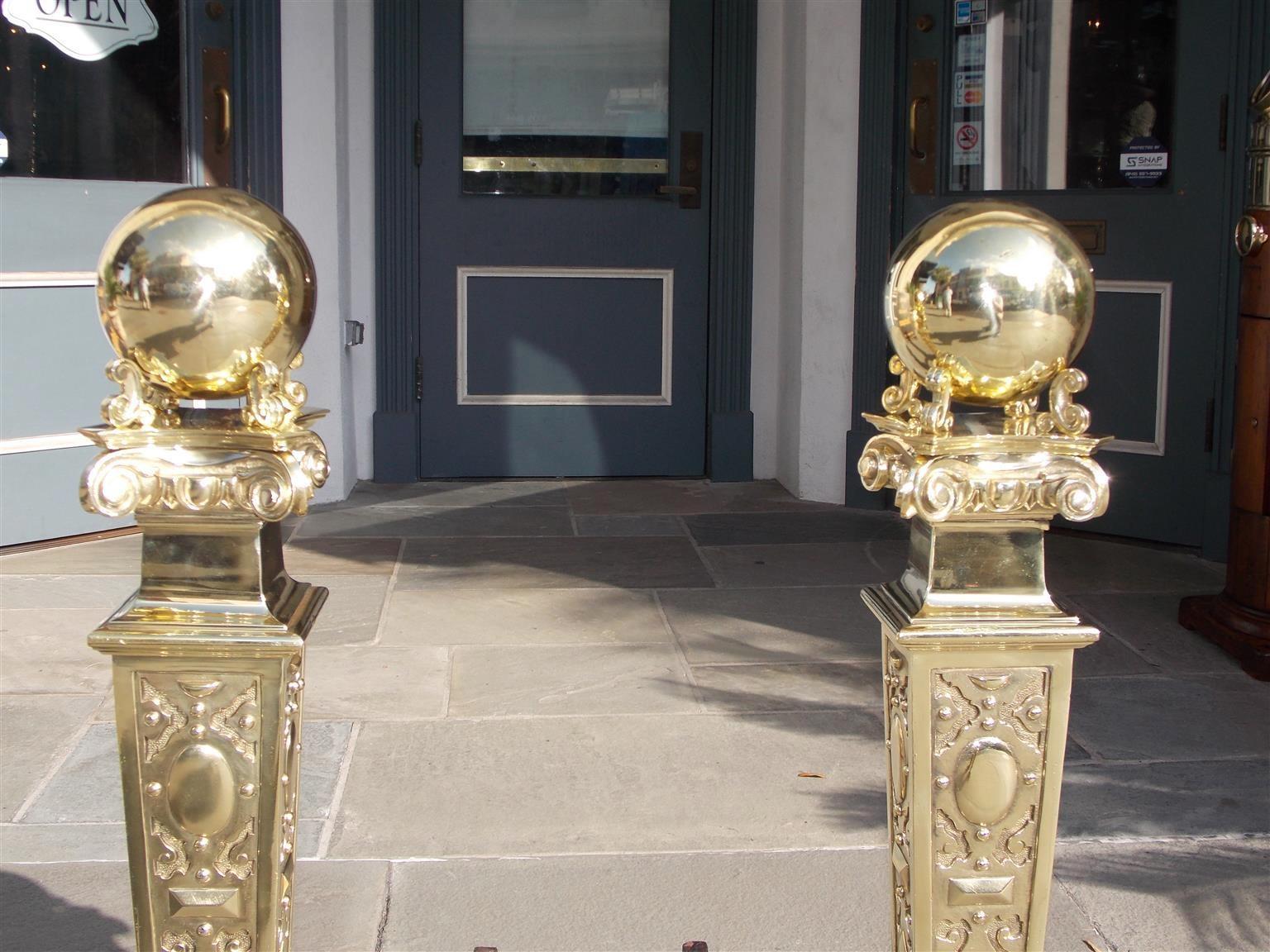 Pair of American Monumental brass andirons with flanking ball finials, Ionic capitals, tapered decorative medallion plinths, spit hooks, matching squared tapered log stops, original wrought iron dog legs, and resting on arched centered medallion
