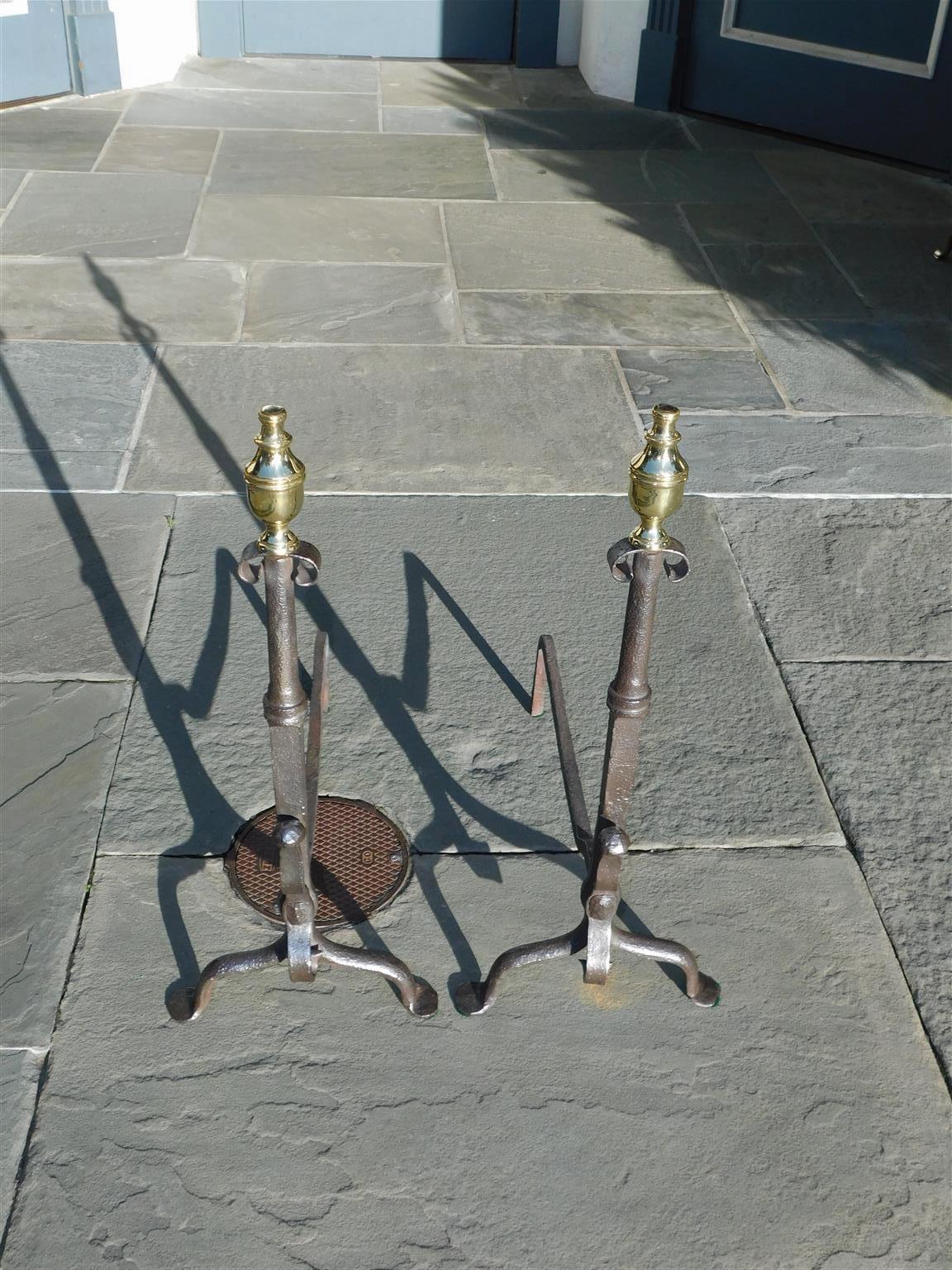 Pair of American wrought iron and brass andirons with flanking urn finials, decorative scroll work, squared circular plinths with spit hooks, matching scrolled legs, and resting on the original penny feet. Late 18th century. Central plinth columns