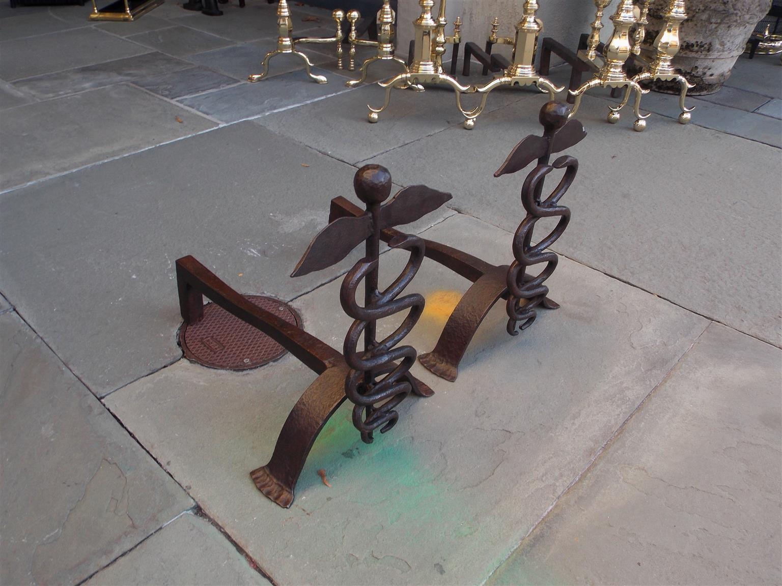 Pair of American wrought iron ball finial caduceus andirons with stylized splayed feet, Mid-19th century.
The caduceus is the traditional symbol of Hermes and features two snakes winding around an often winged staff and is often used as a symbol of