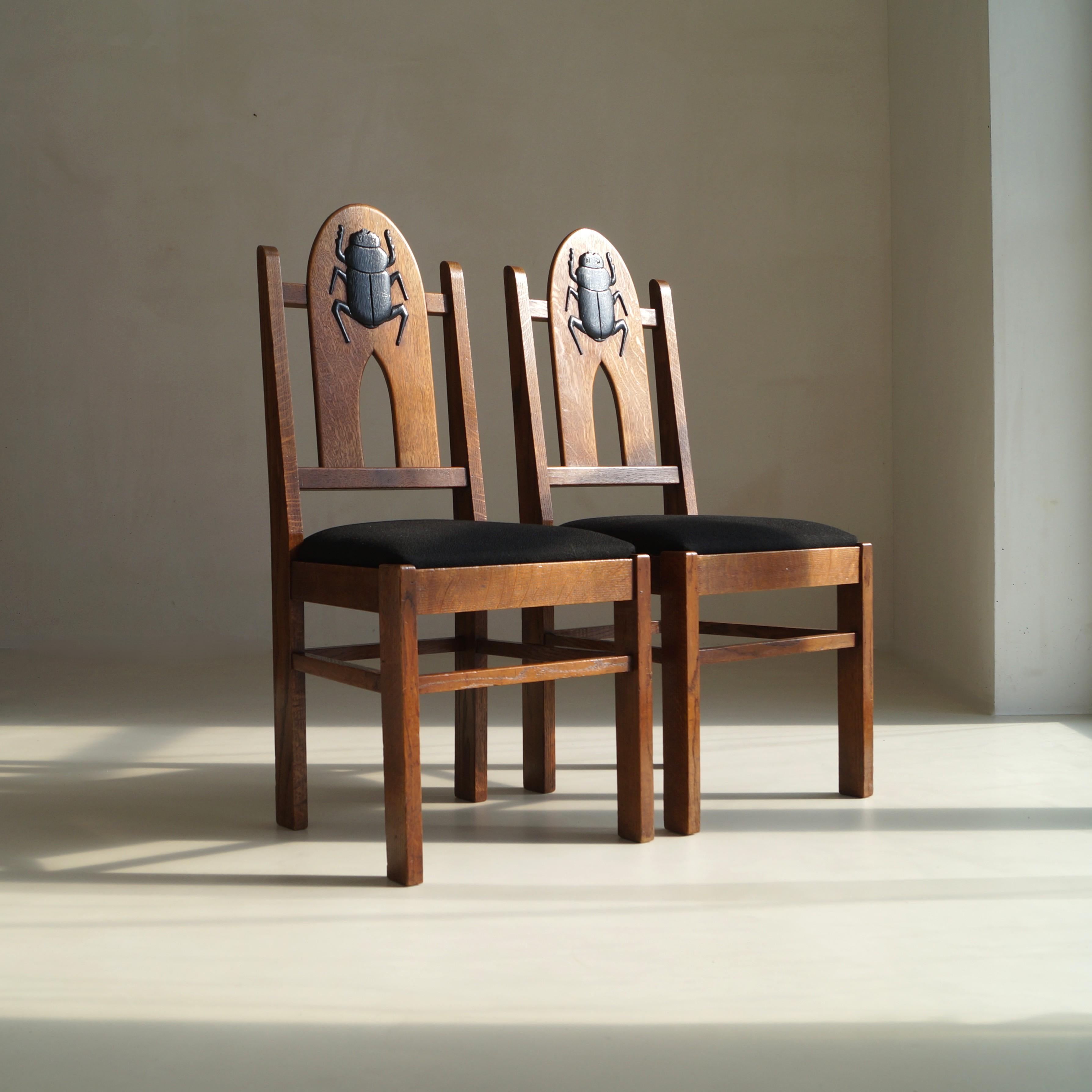 A stunning and rare pair of Dutch Art Deco Amsterdam School chairs dating from c. 1925, beautifully crafted in solid oak with upholstered black seats. Each chair features a hand-carved scarab beetle on the backrest - a clear nod to the Egyptomania