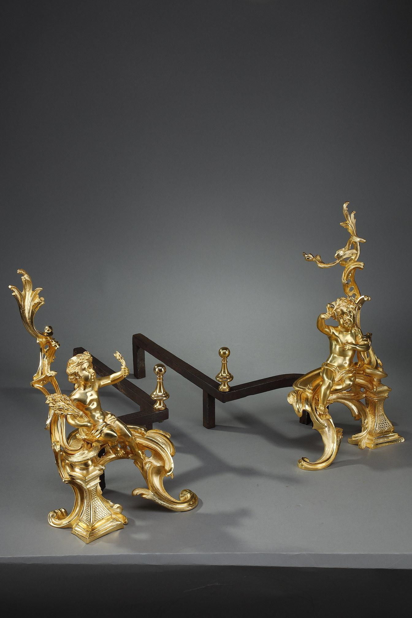 Pair of Louis XV style andirons with rocaille decoration. They are decorated with ormolu and chased putti sitting on a moving base. One putto wearing a crown of wheat is holding a sheaf of wheat, while the other holding his drape is holding flowers.