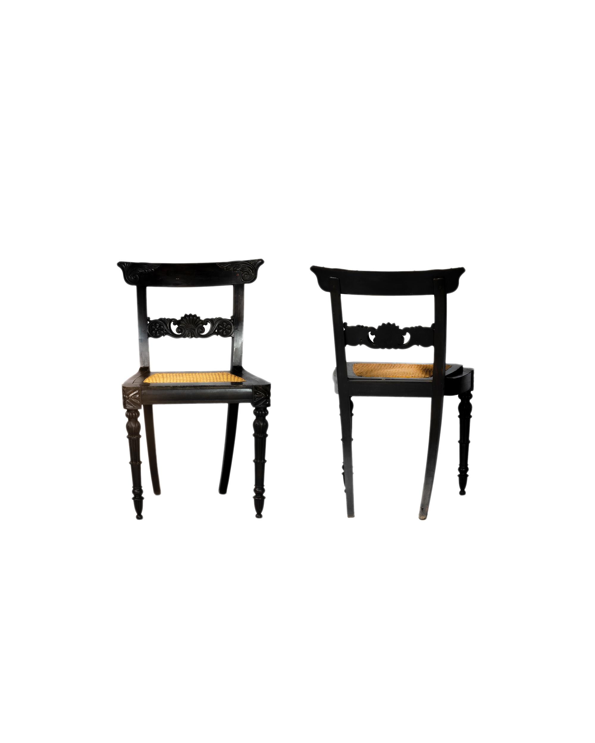 A fine set of two Anglo-Indian Ceylonese solid ebony chairs, horizontal splats with floral motifs standing on reeded front legs and plain back and caned seats. 
The chairs' reeded frames add texture, the horizontal splats showcase intricate floral