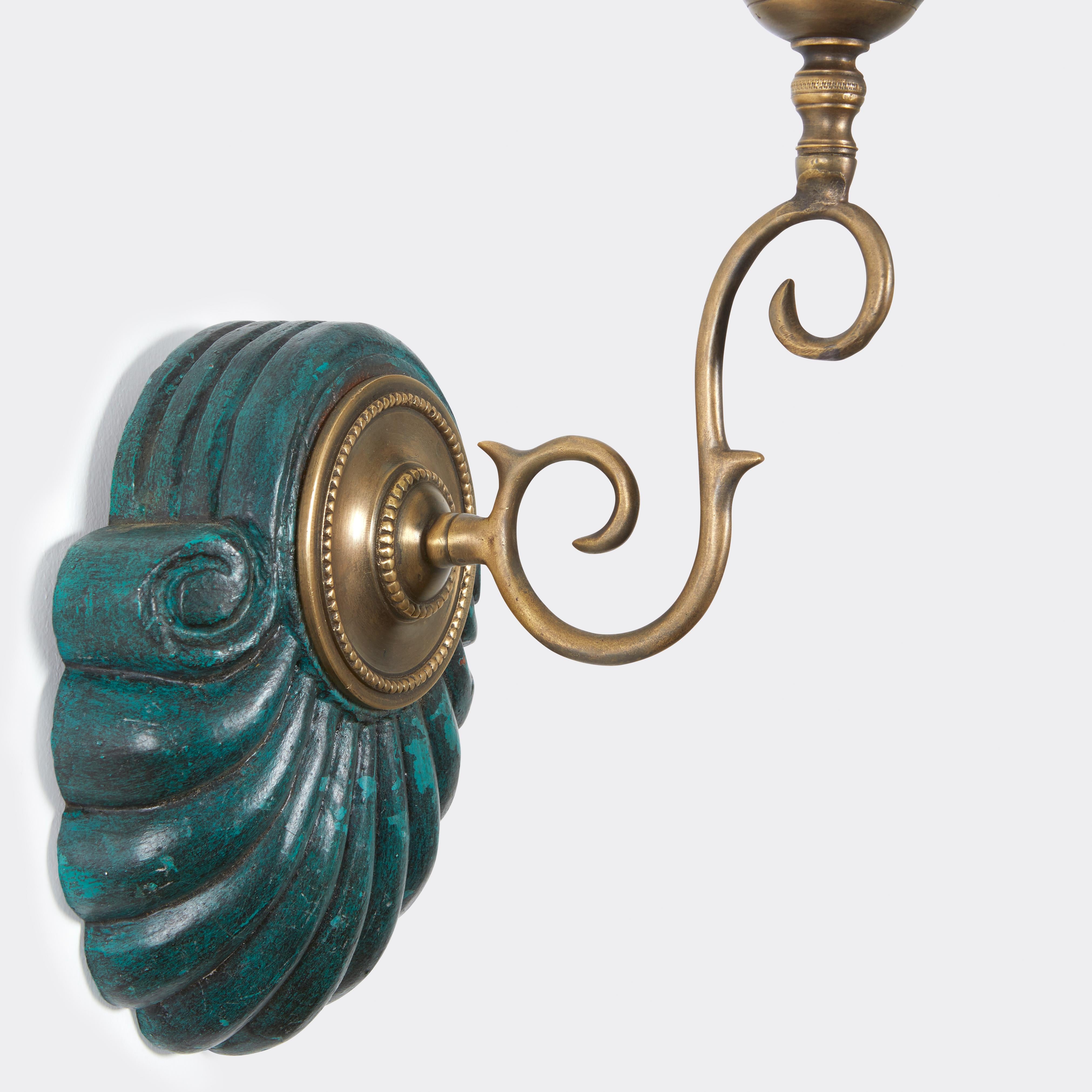 A pair of hand-carved shell form Anglo Indian backplates with a verdigris painted surface, brass sconce fittings and light green hurricane shades. Sconce is measured from bottom of backplate to top of shade height 23